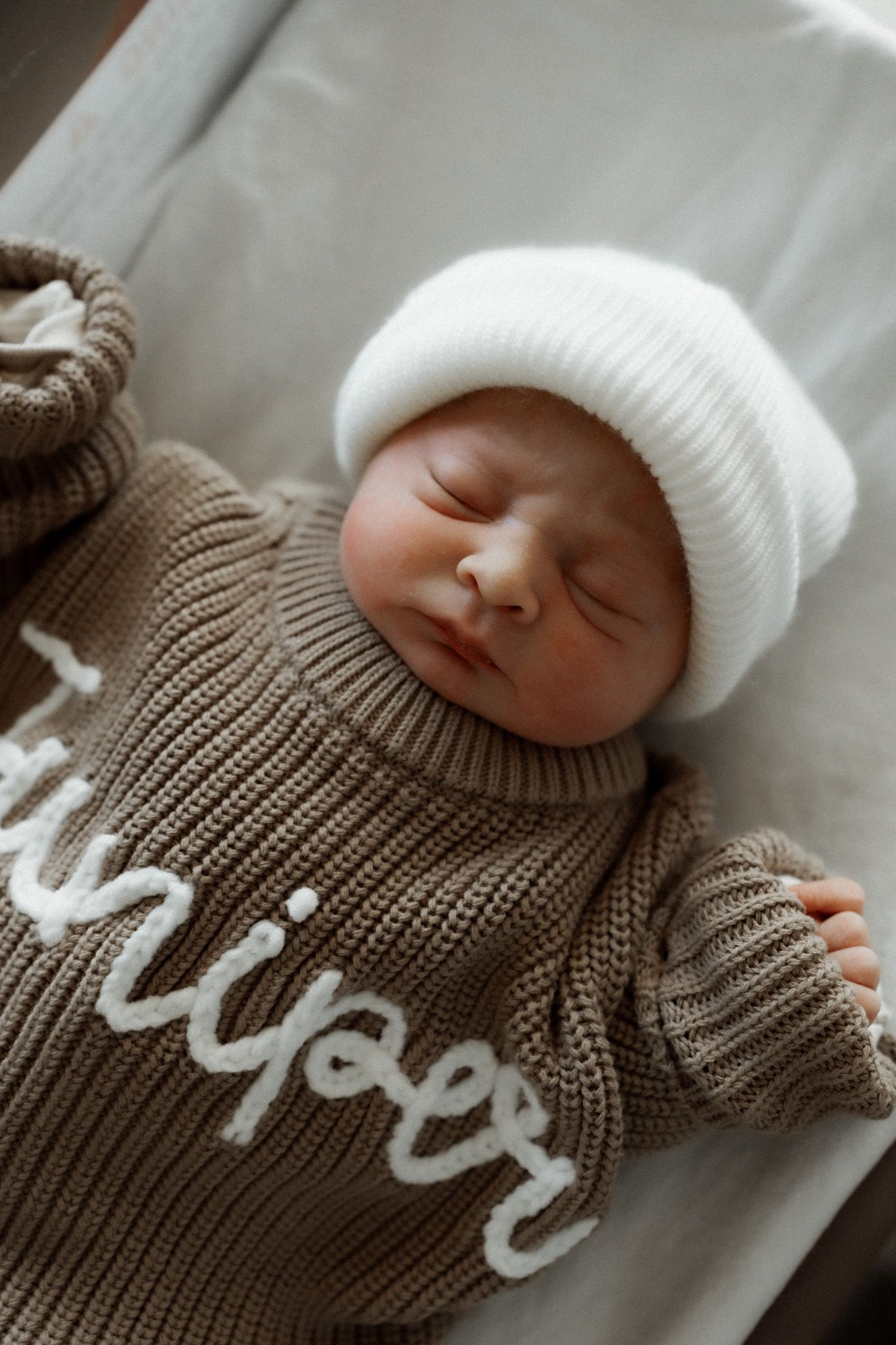 Newborn baby wearing white beanie and brown knit sweater sleeping on white sheets.