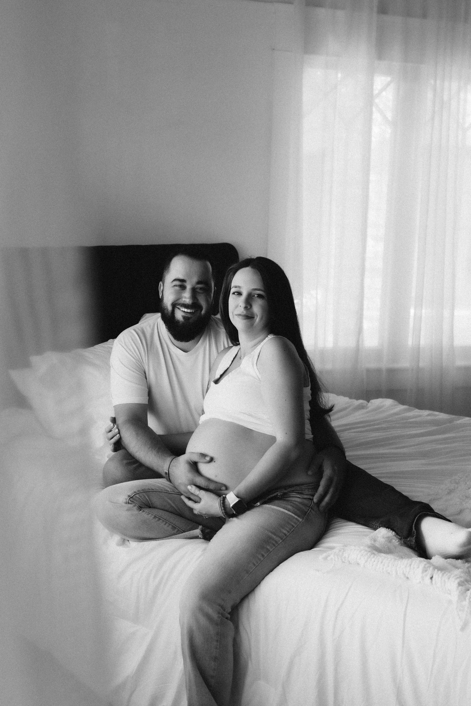 Black and white maternity photo of couple sitting together on bed.