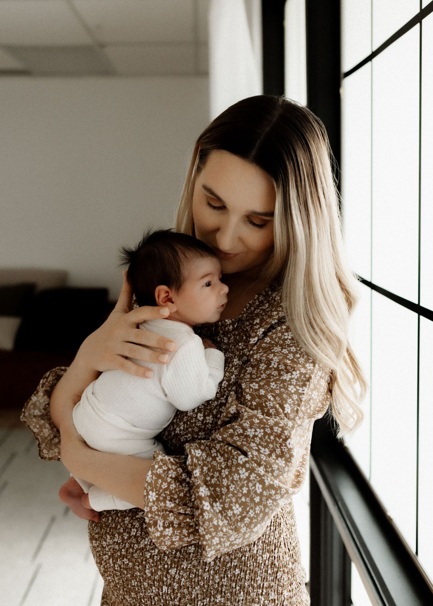 Parent in floral dress holding baby near window in sunlit room.