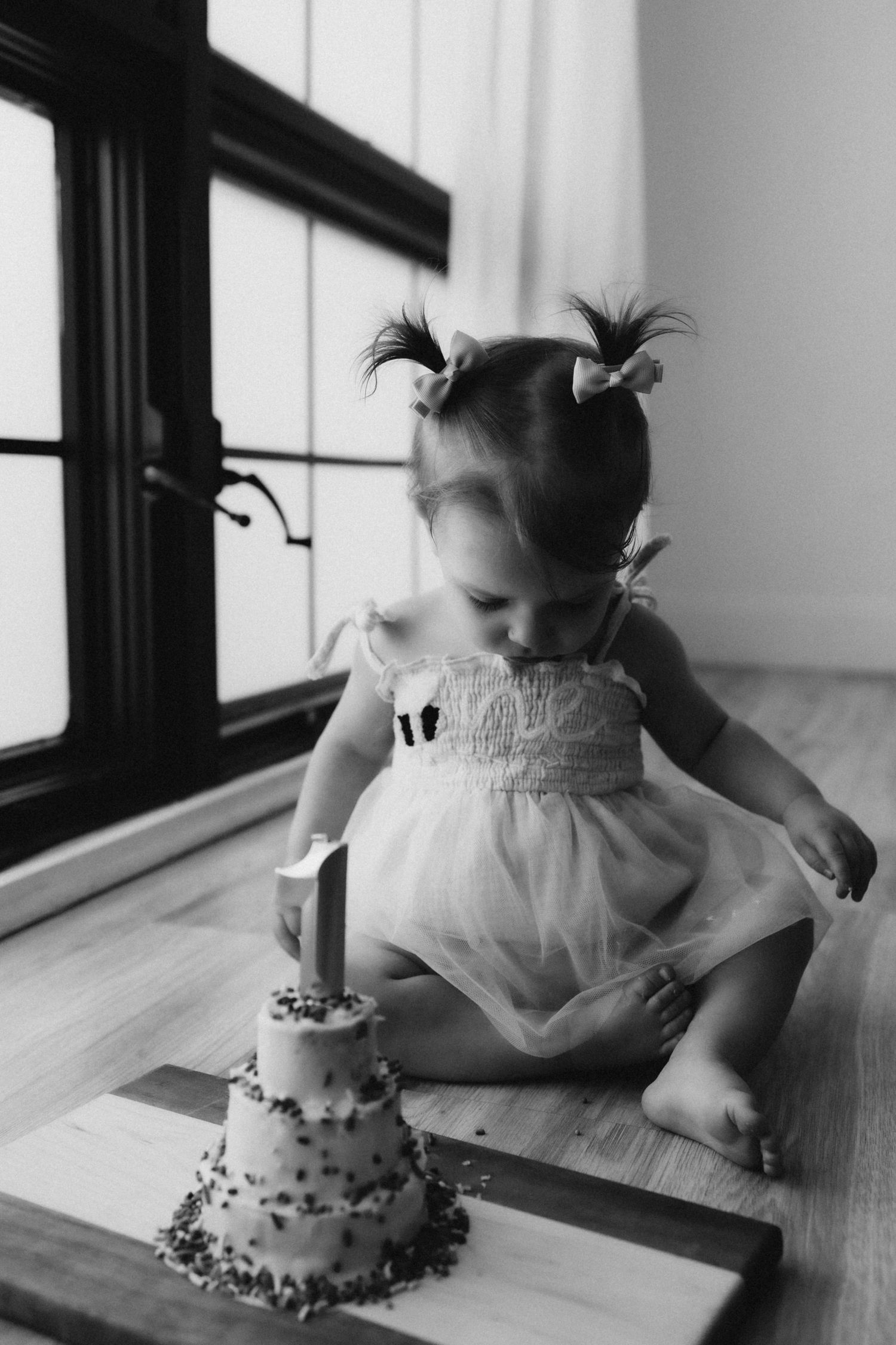 Black and white photo of small toddler in white dress looking down at tiny birthday cake on wooden floor.