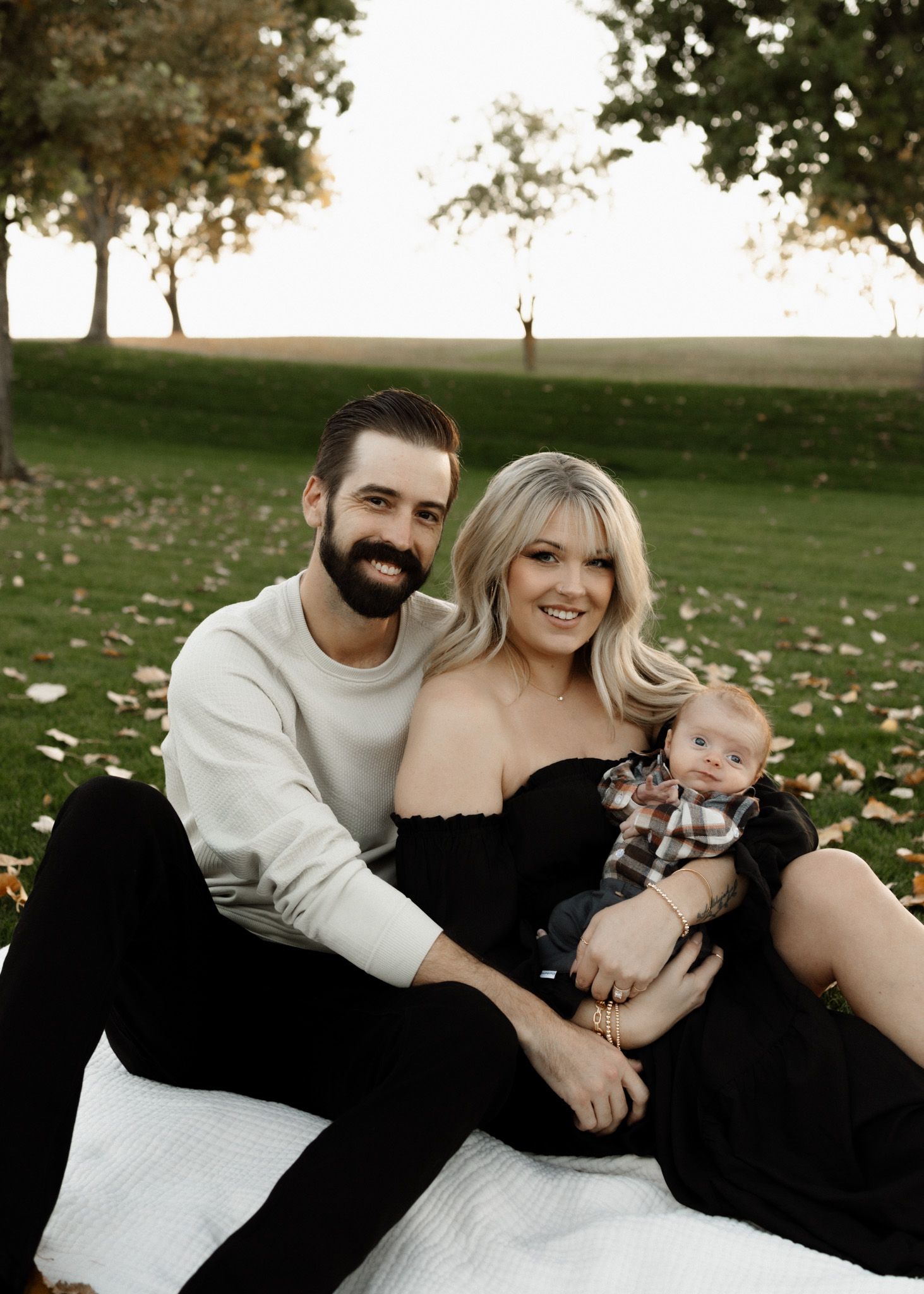 Family portrait on white blanket with autumn trees and scattered leaves in background.
