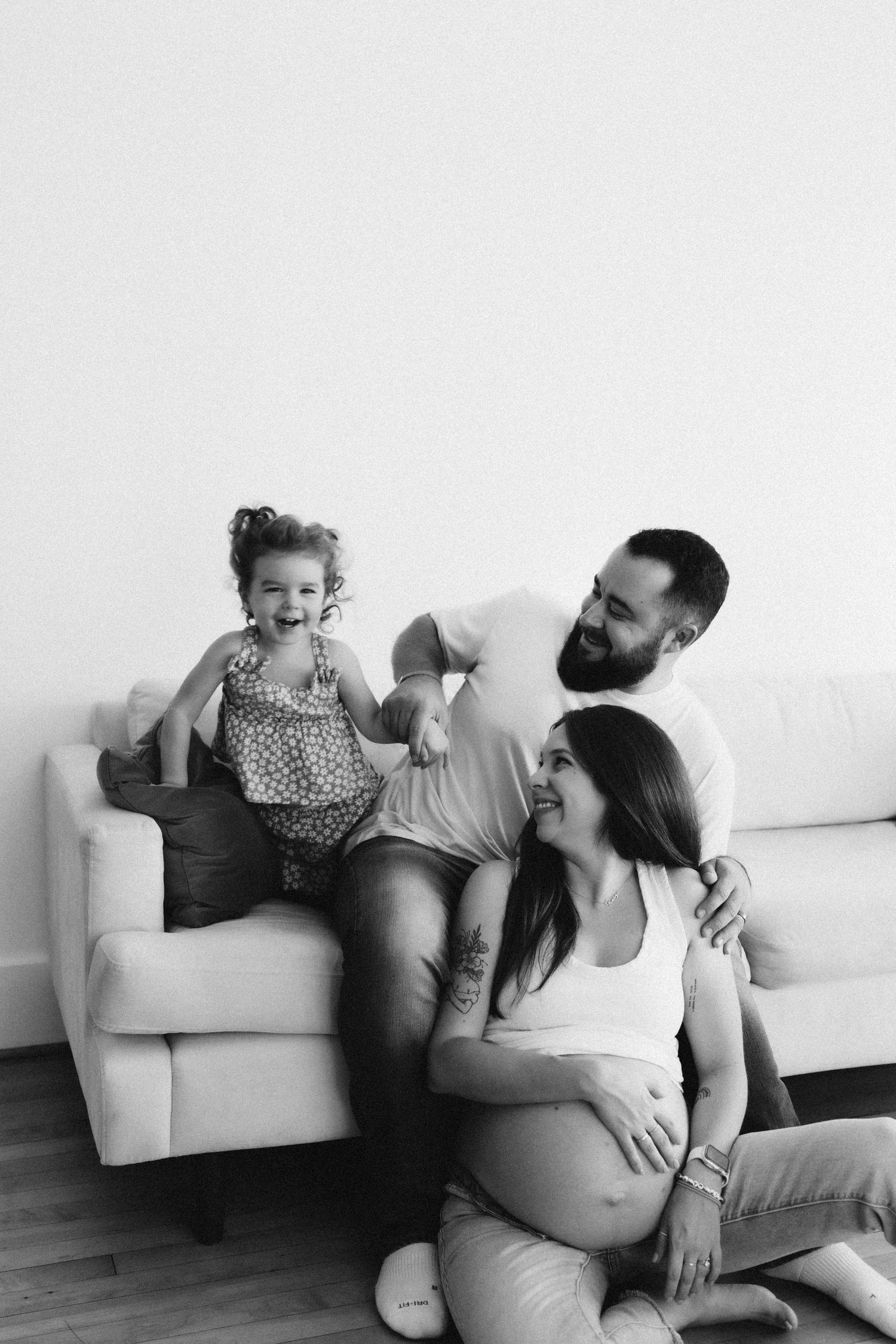 Black and white candid photo of family lounging on white couch showing playful interaction.