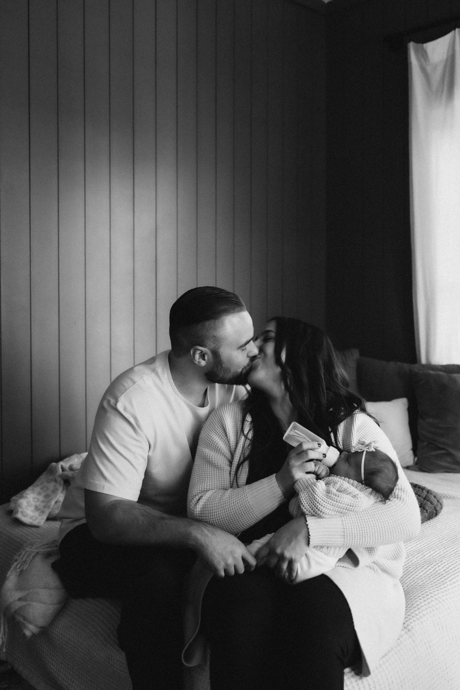 Intimate black and white photo of parents sharing a loving moment with their newborn on a bed.