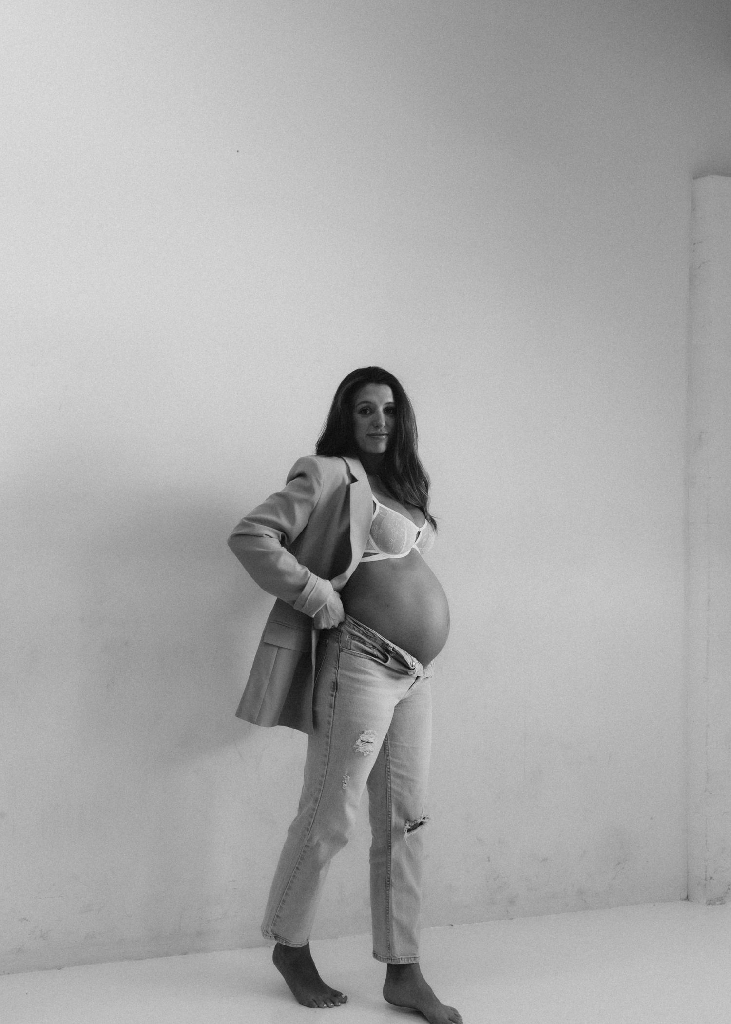 A black and white portrait of someone in a casual blazer and jeans showing off their pregnant belly against a white wall.