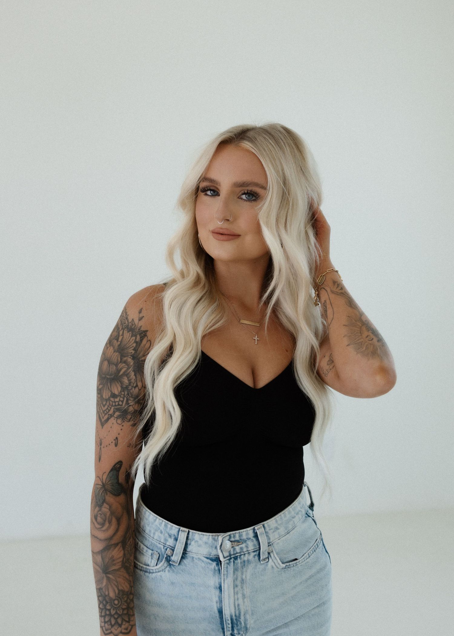 Woman in black tank top and light wash jeans poses against white background showing sleeve tattoos on both arms.