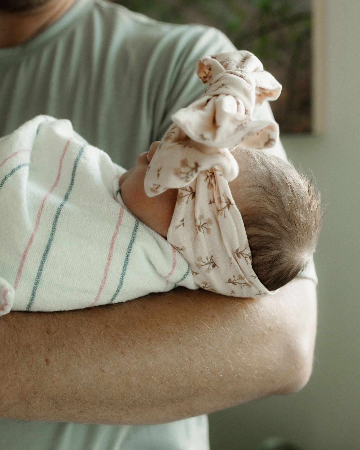 A newborn baby swaddled in white cloth with a floral headband rests peacefully in someone's arms.