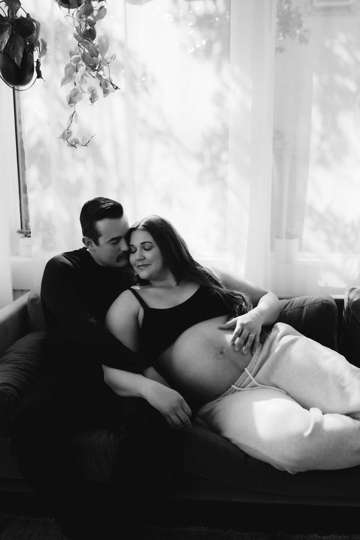 A tender maternity photo of an expectant couple lounging together on a dark couch in black and white.