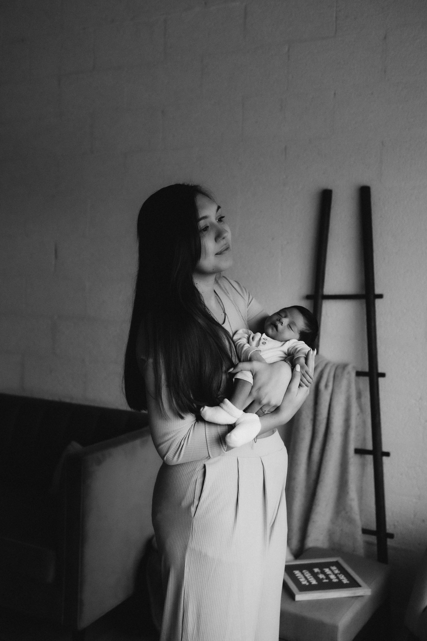 Black and white portrait of nurturing moment with sleeping infant against brick wall backdrop.