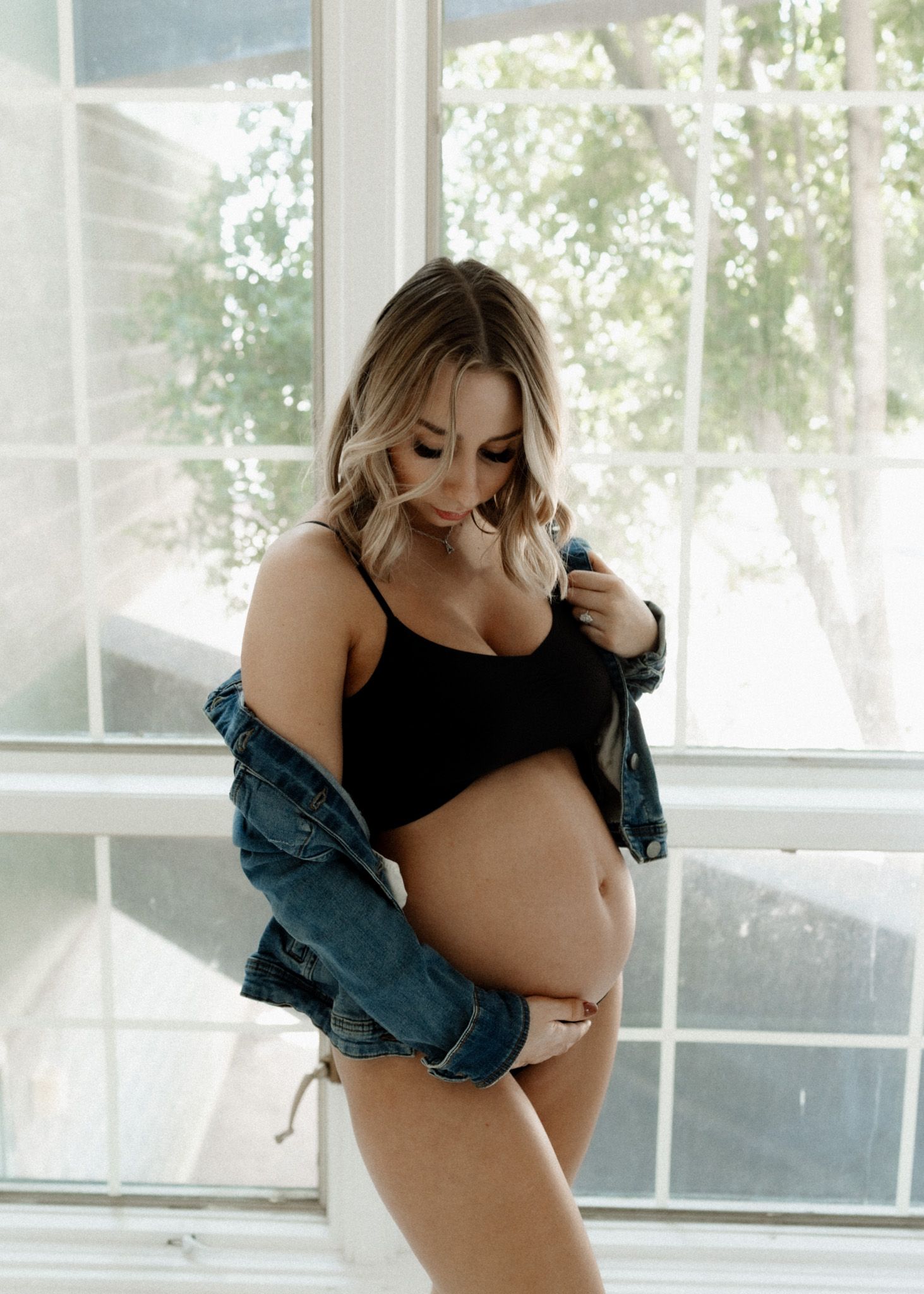 Maternity photo in denim jacket showing pregnant belly near bright windows.
