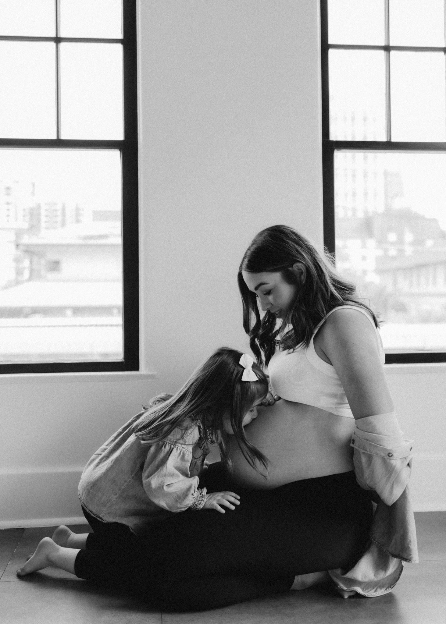 A tender black and white maternity moment between mother and child by a window.