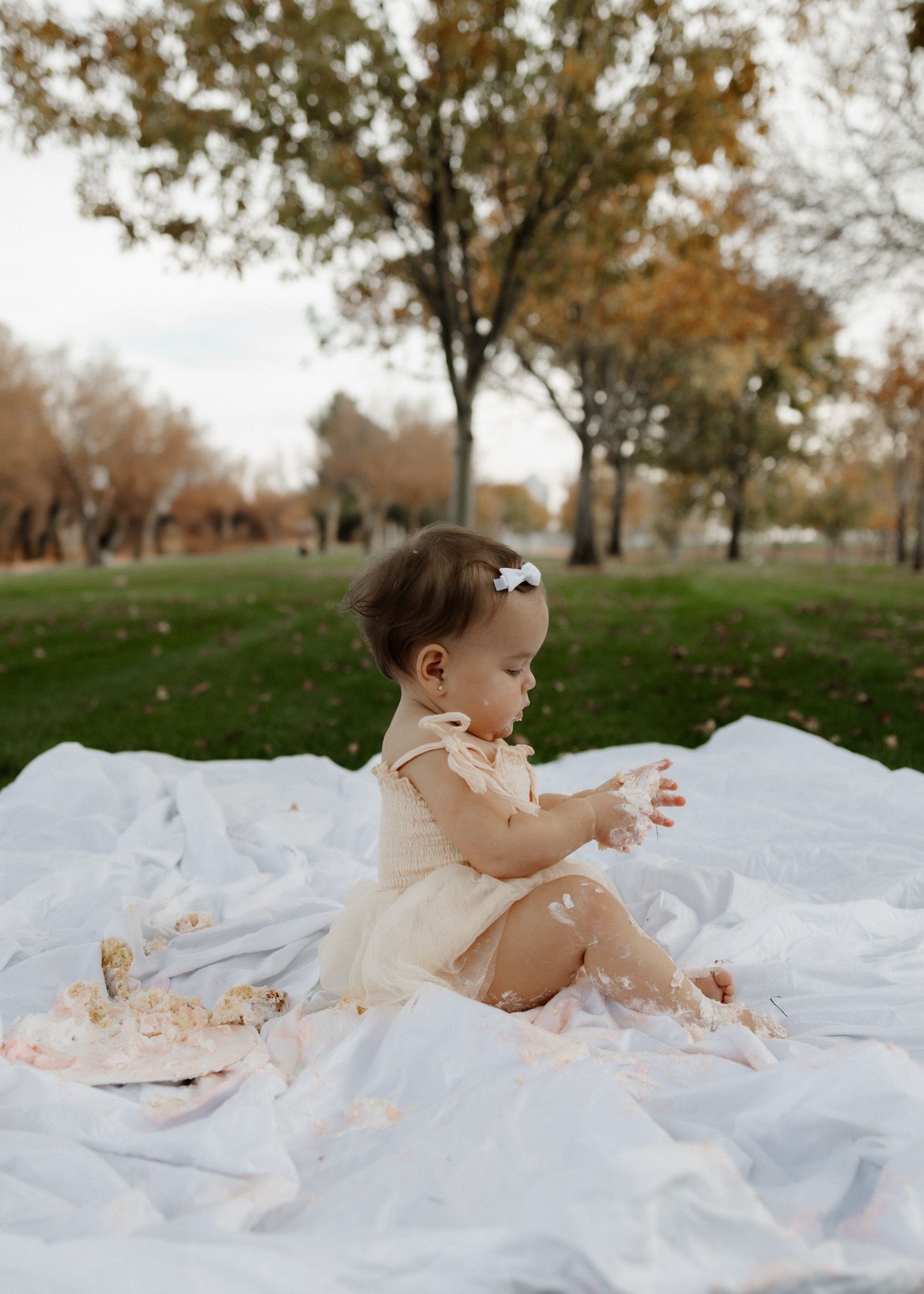 A baby in cream dress sits among cake crumbs during outdoor first birthday photoshoot.
