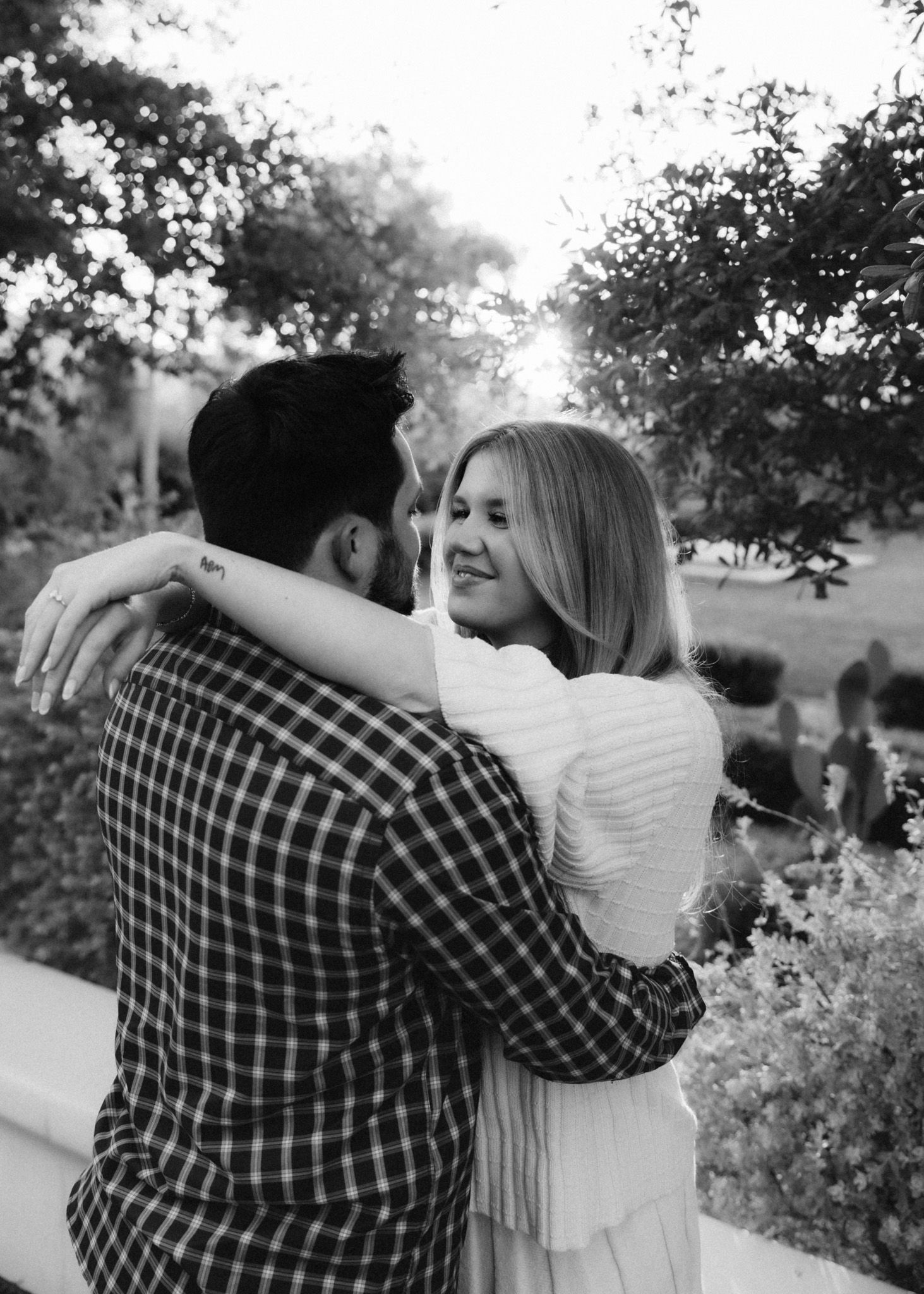 Black and white photo of a couple embracing outdoors with trees in background.