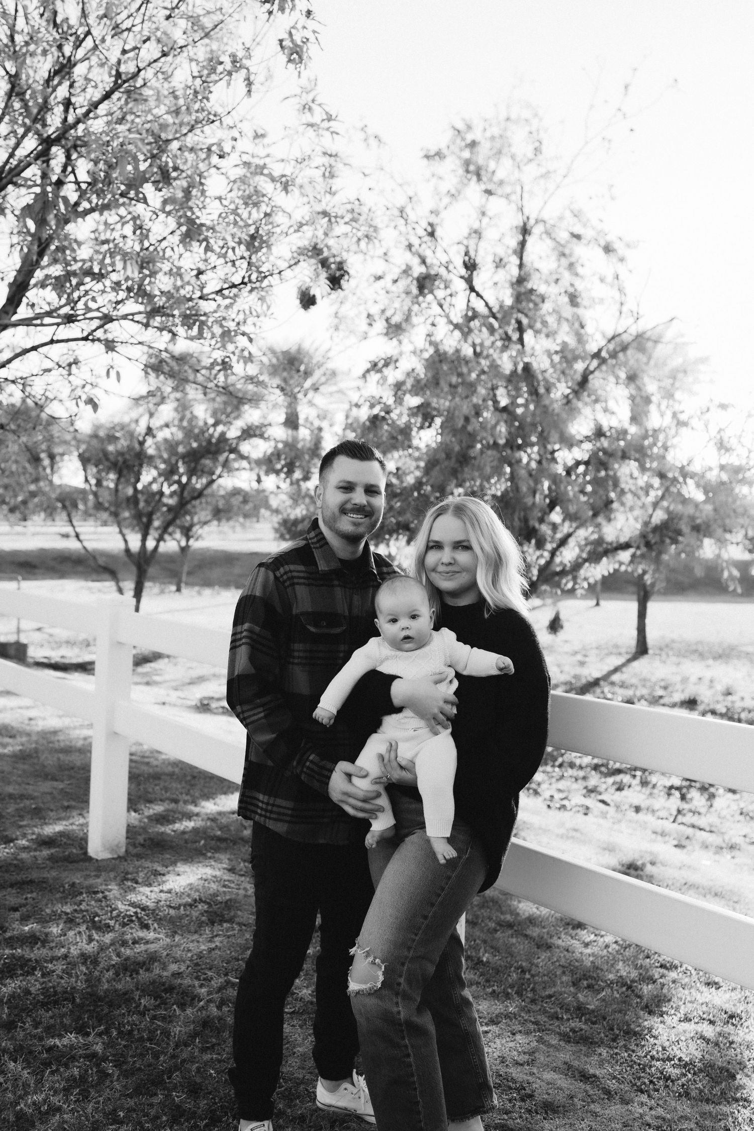 Family posing together by a white fence with trees in the background.