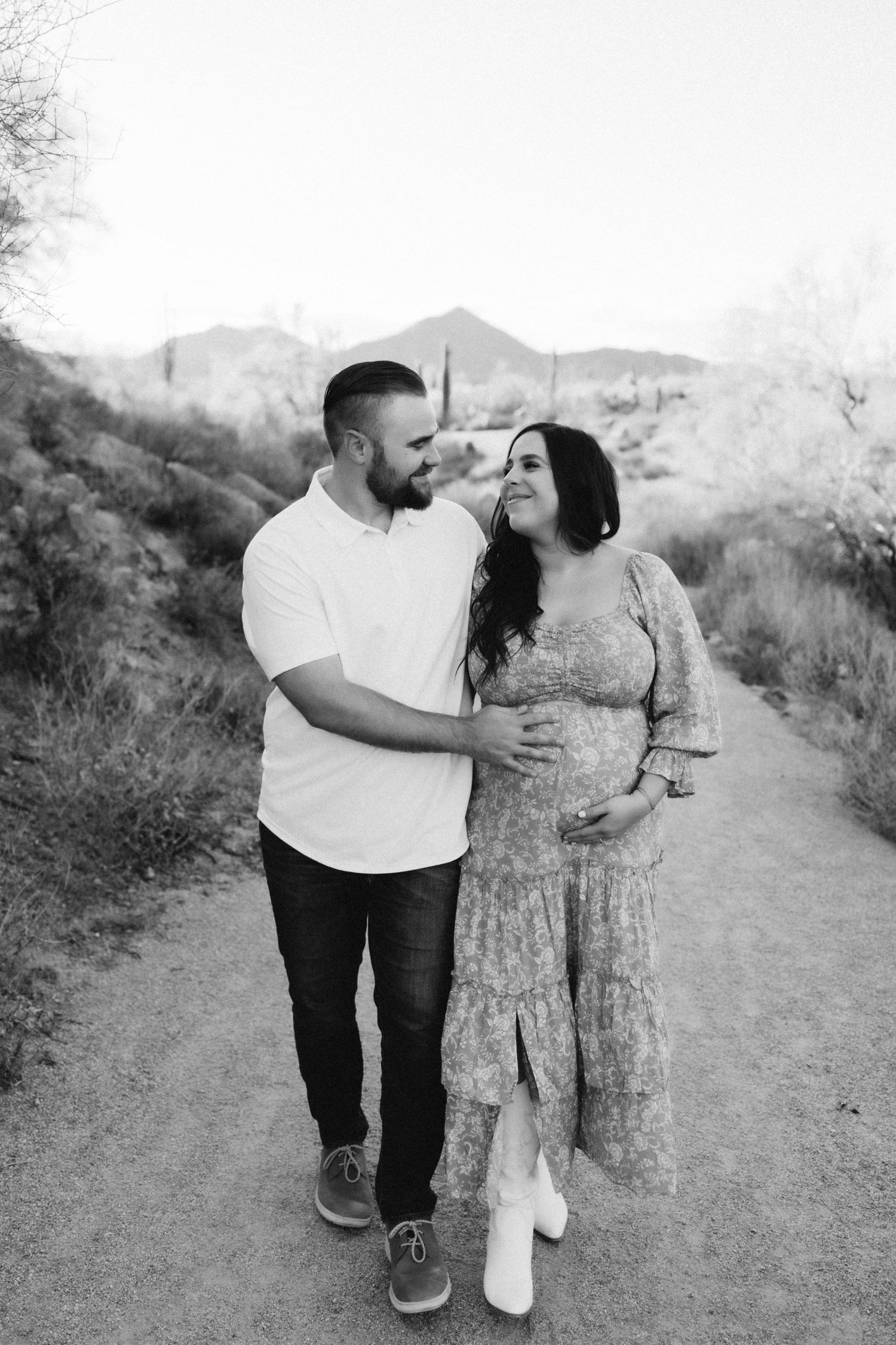 A couple walking on a desert trail during a maternity photo session against mountain backdrop.