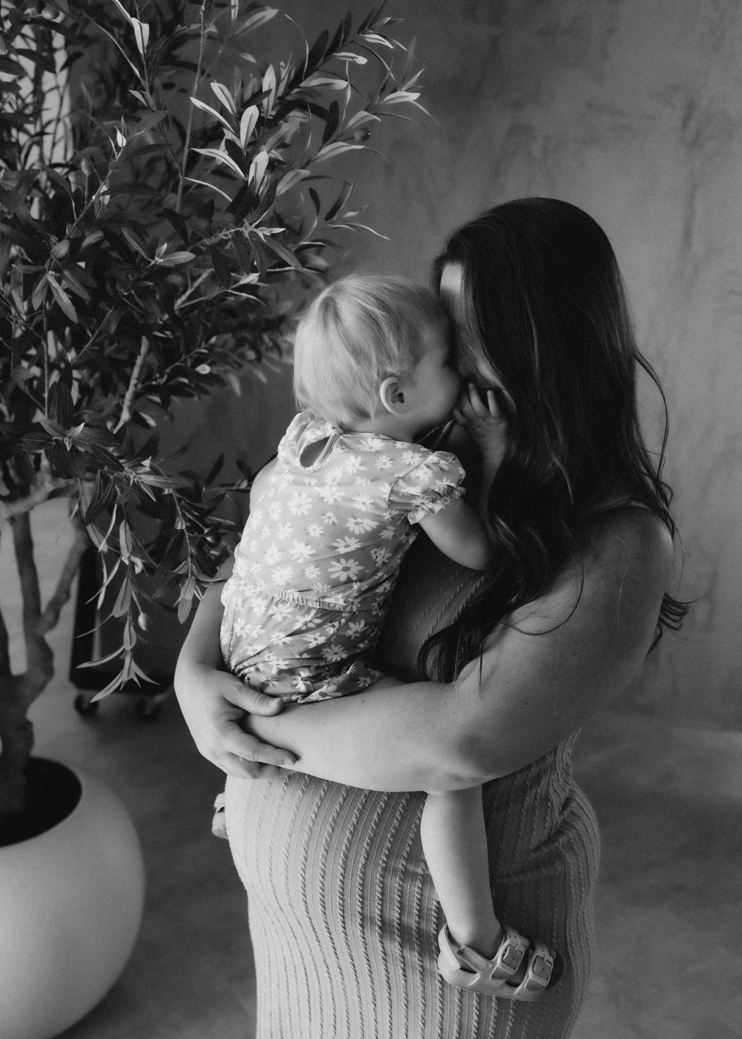 Mother holding a toddler next to a potted plant in a black and white portrait.