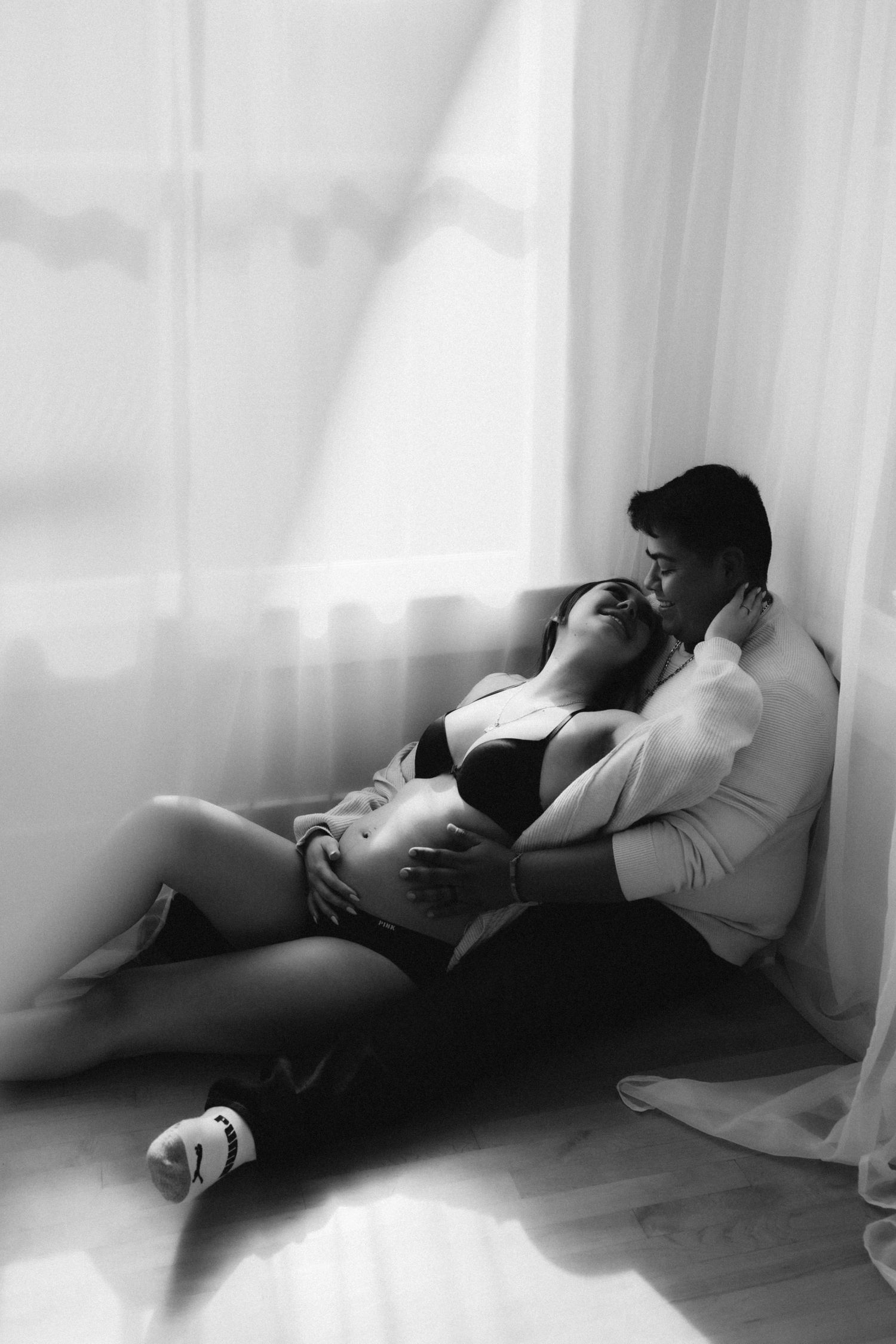 A black and white intimate maternity photo of a couple resting together by a window.