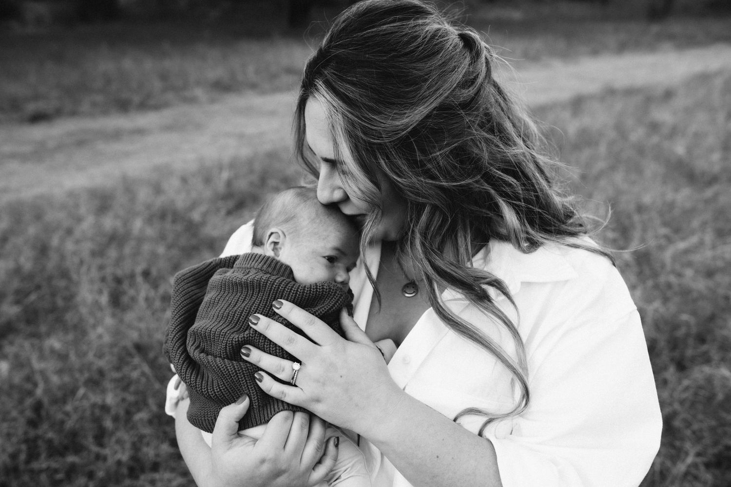 Black and white photo showing tender moment between parent and baby in outdoor field setting.