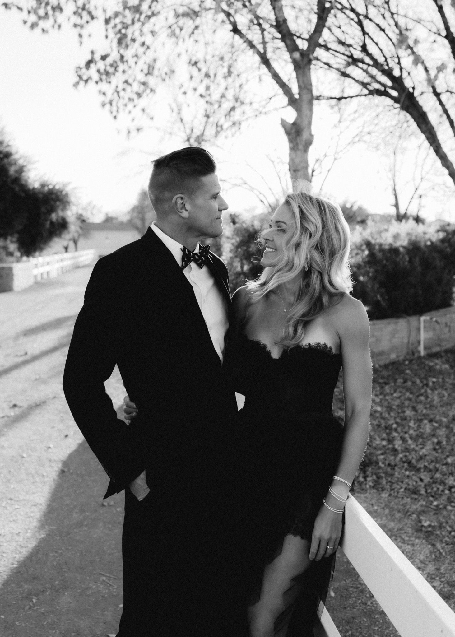 Elegant couple in formal attire pose together on a tree-lined path in black and white.