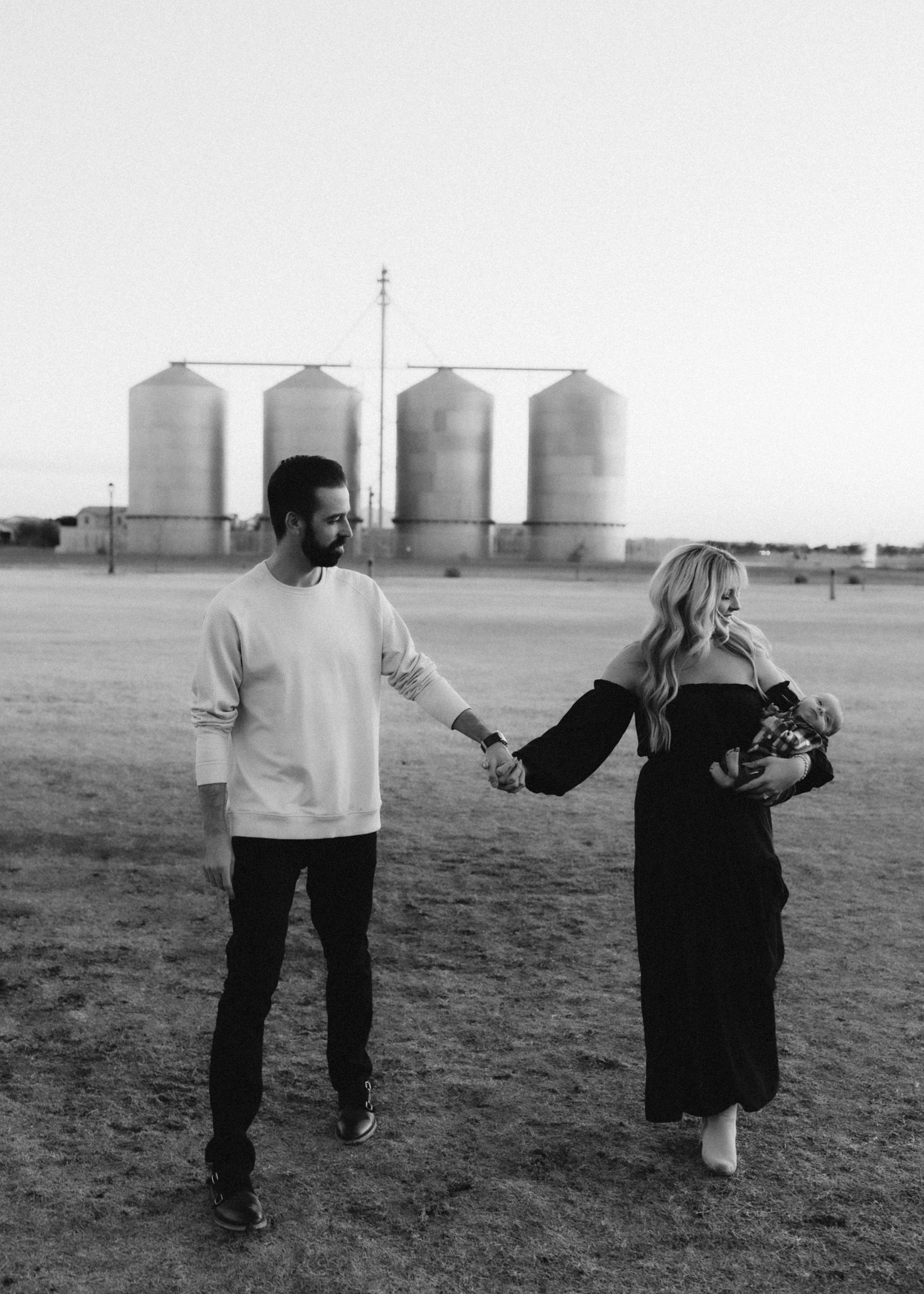Black and white photo of a couple holding hands in front of grain silos in an open field.