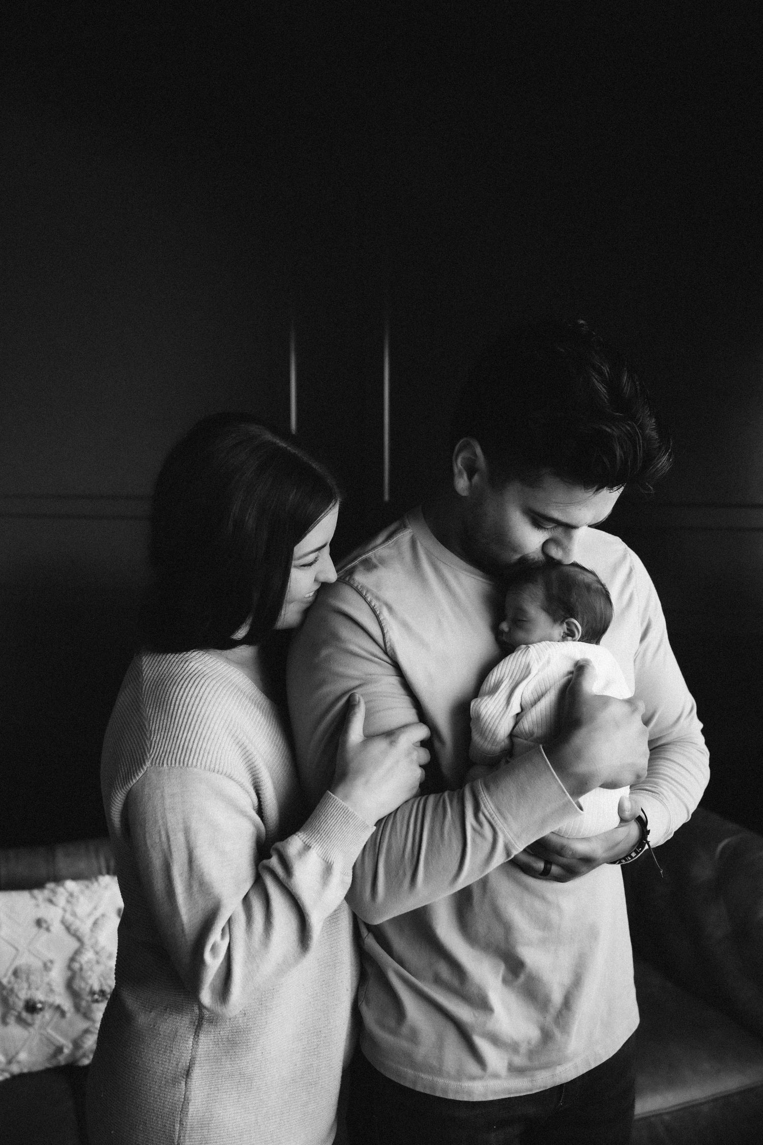Black and white portrait of parents tenderly holding their newborn baby against a dark wall.