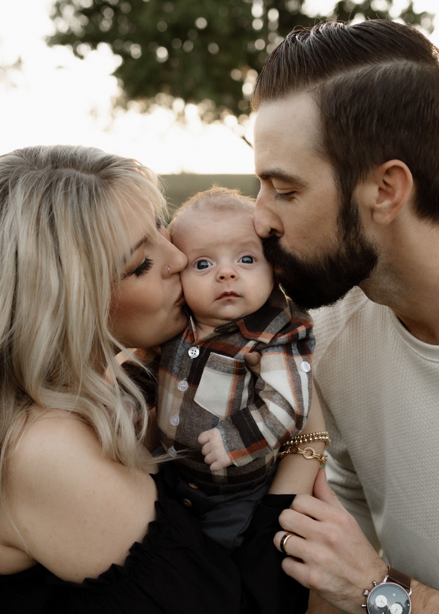 Parents kissing baby on cheeks in outdoor autumn setting.
