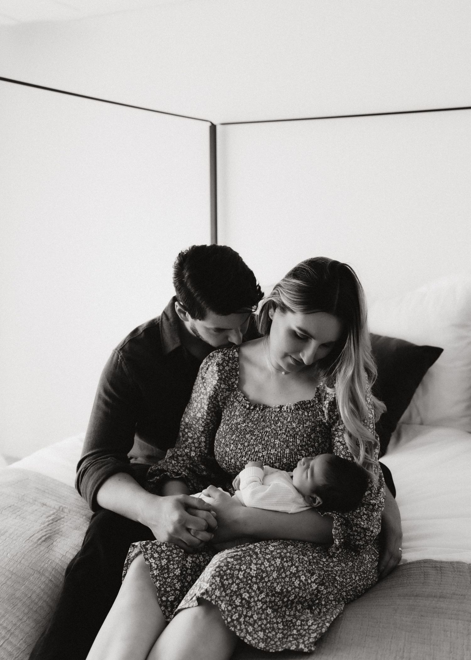 An intimate black and white portrait of new parents cradling their newborn baby on a bed.