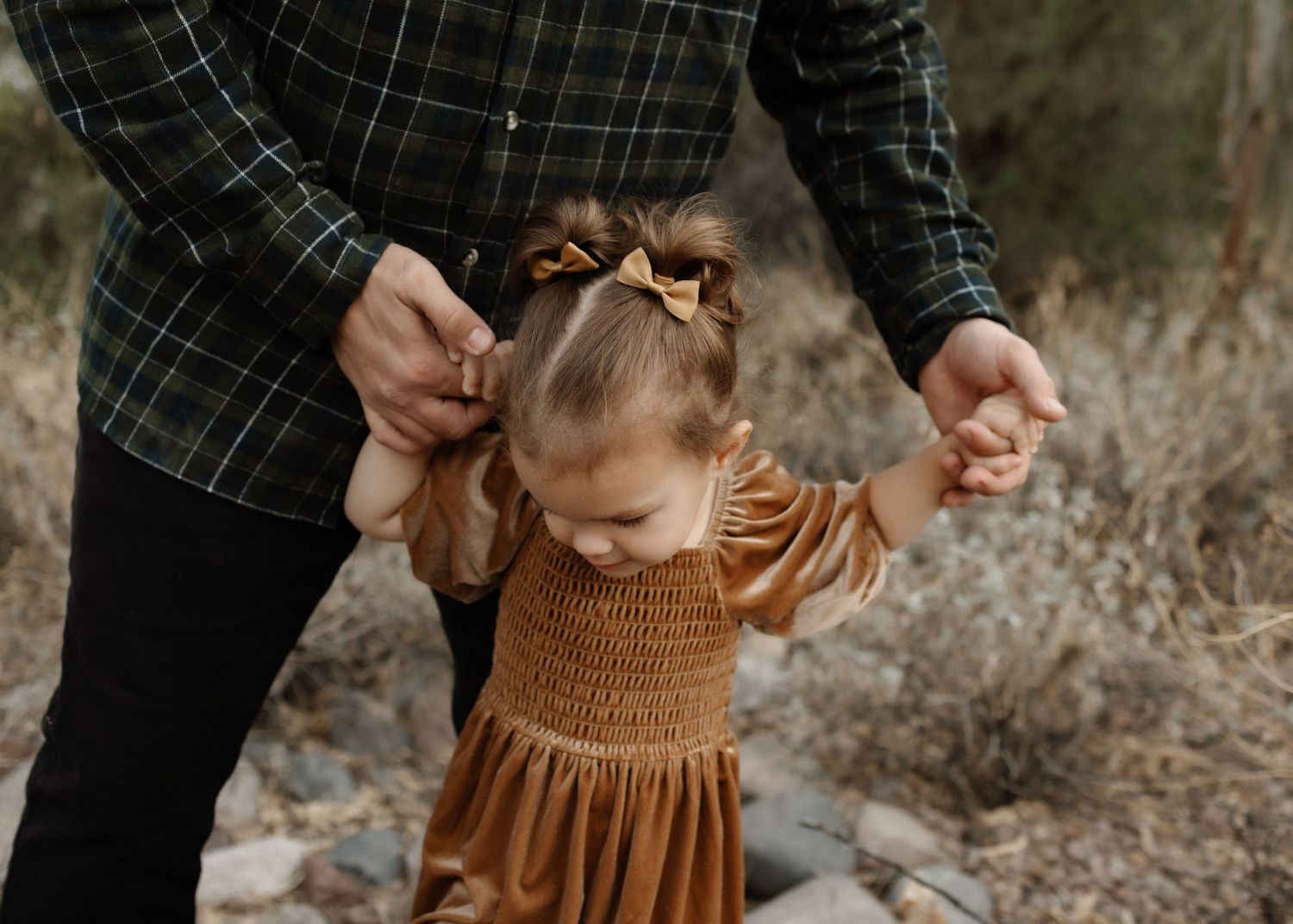 Small child wearing a brown smocked dress holds hands with someone in a plaid shirt in an outdoor desert setting.