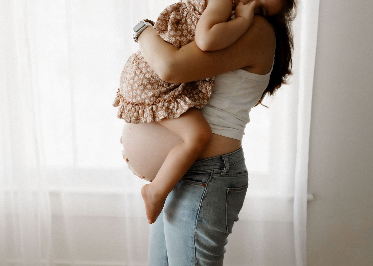 Pregnant woman in jeans holding toddler against white curtain backdrop.