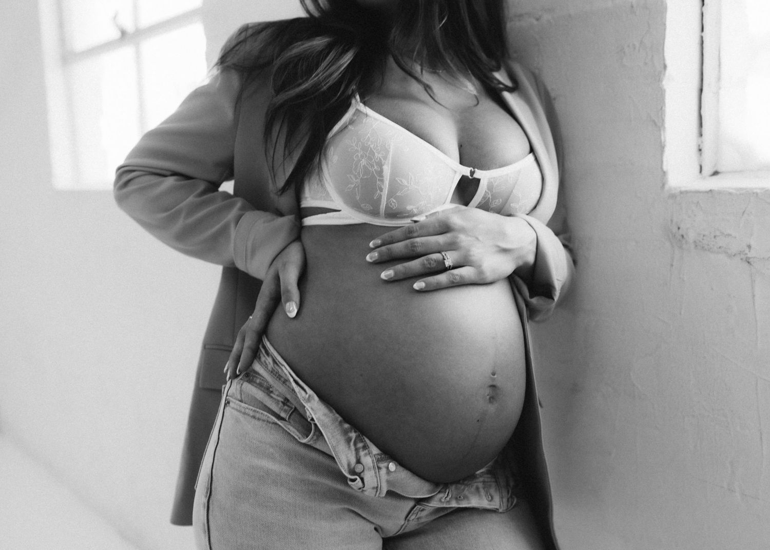 Black and white photo of a pregnant belly being cradled by hands, wearing partially unbuttoned jeans.