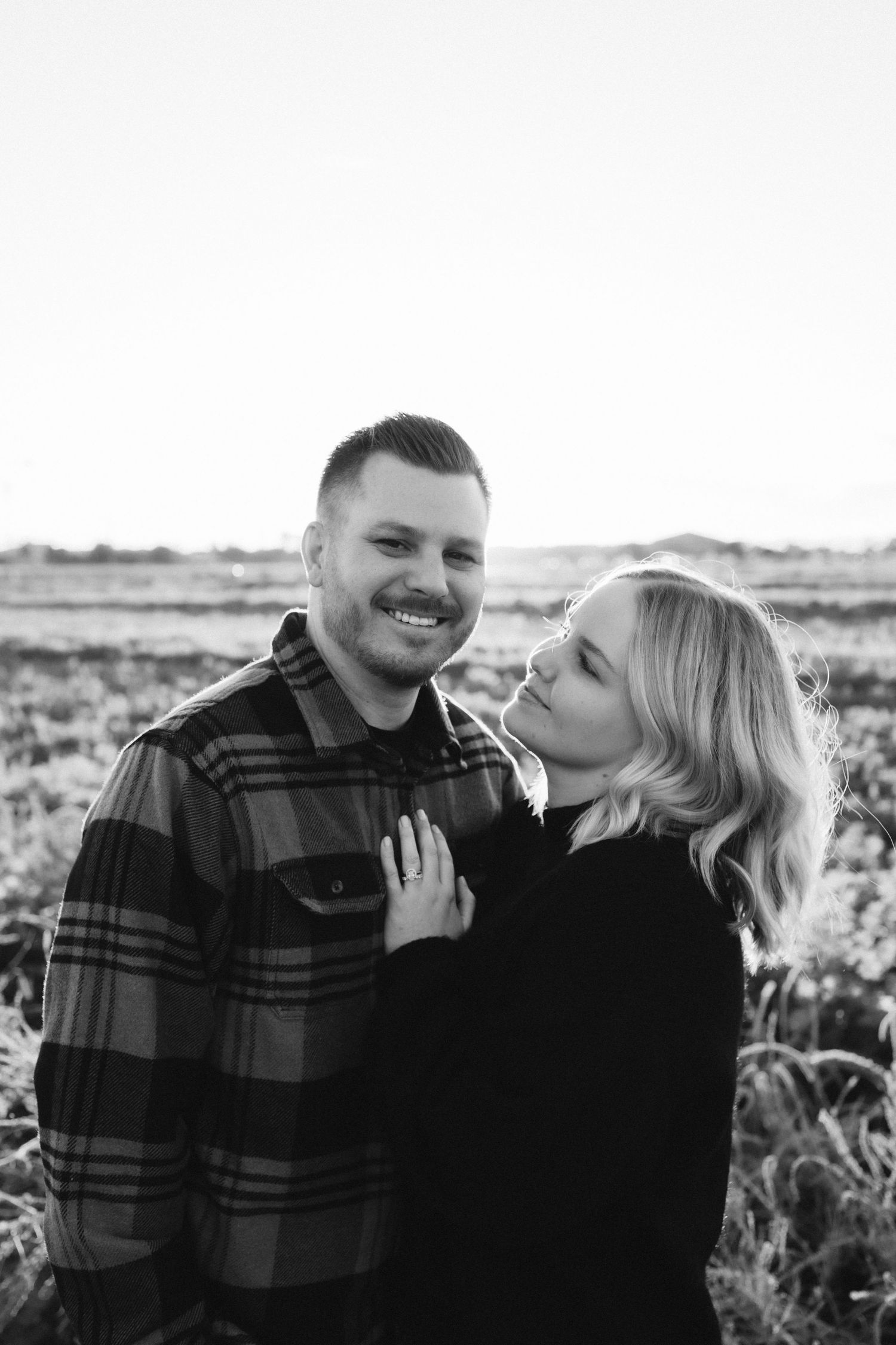 A couple embraces outdoors in a black and white portrait against a natural landscape.