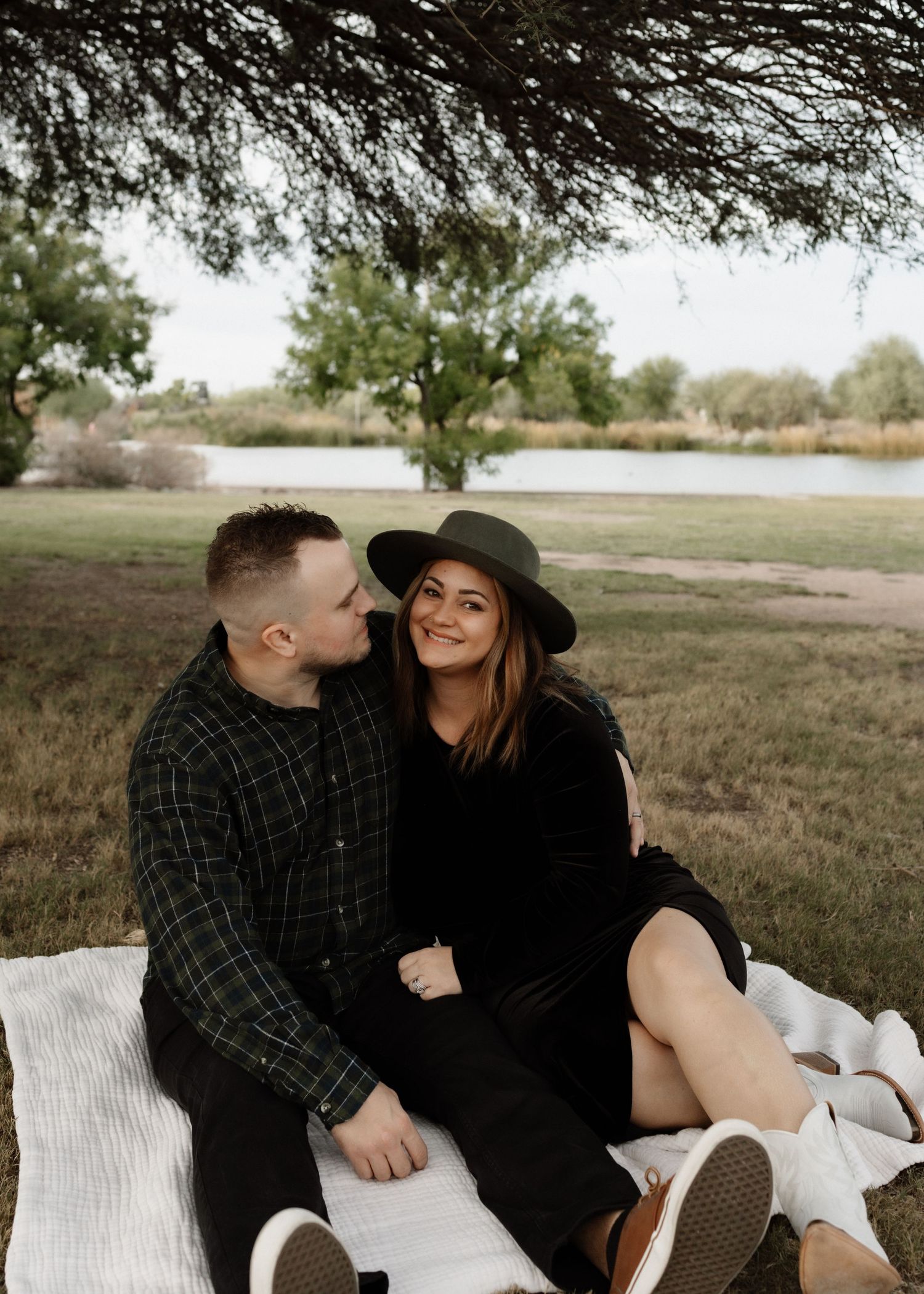 A couple shares an intimate moment while sitting on a blanket by a lake.