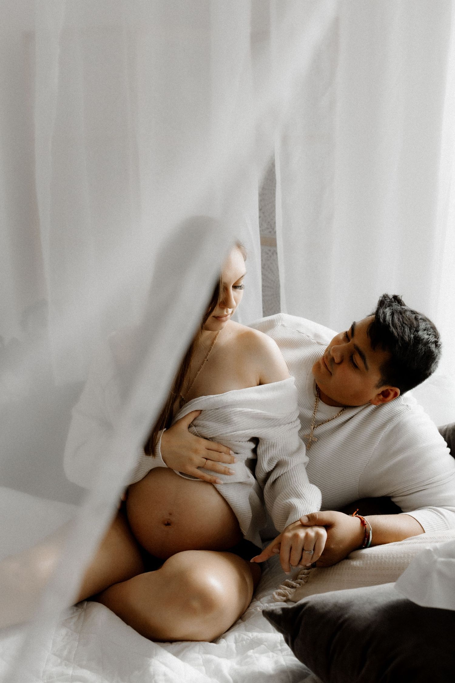 An intimate indoor maternity photo with soft white curtains and natural window lighting.