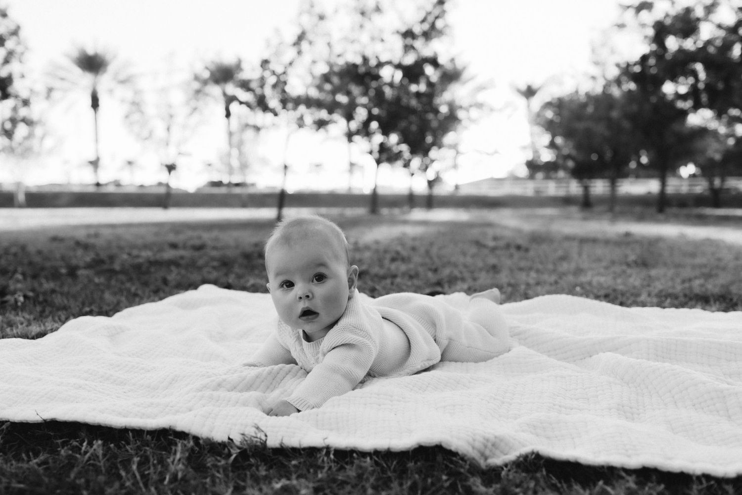 Baby lying on blanket outdoors with windmill visible in background.