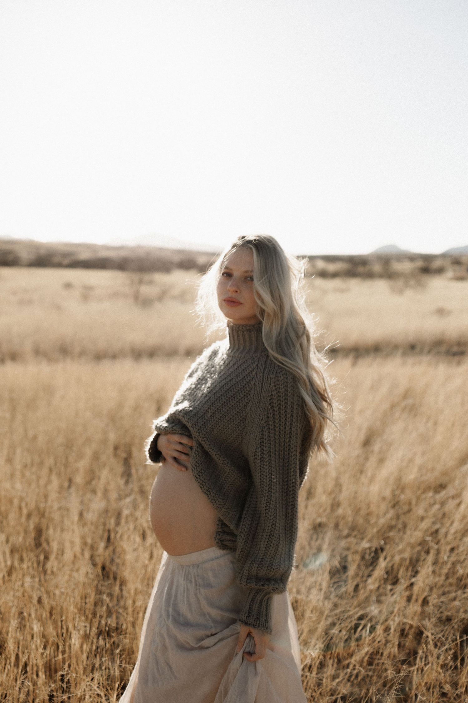 A pregnant woman in a cozy knit sweater stands thoughtfully in a field of golden grass at sunset.