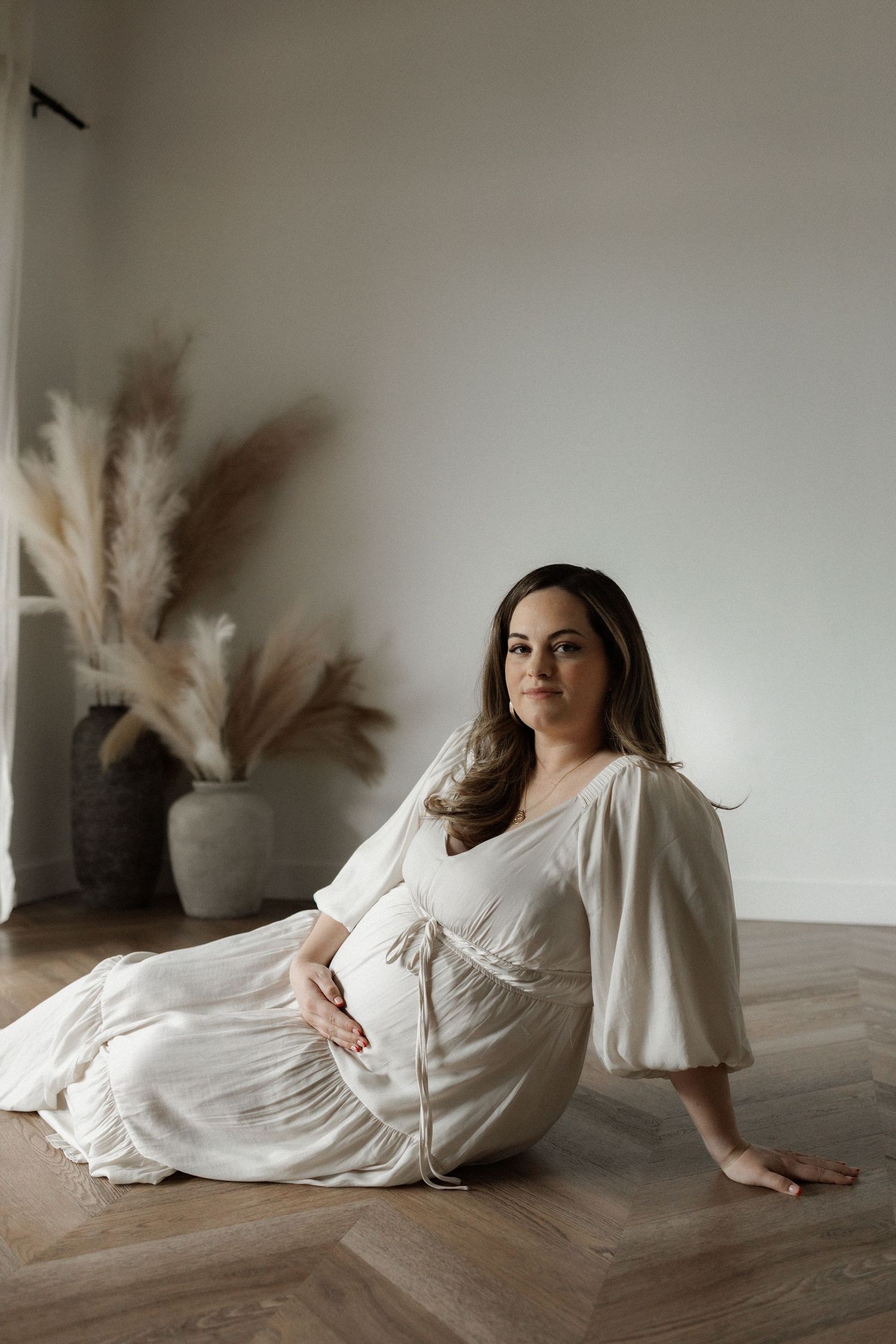 Maternity photo of someone in a flowing white dress sitting on hardwood floor with pampas grass decor in background.