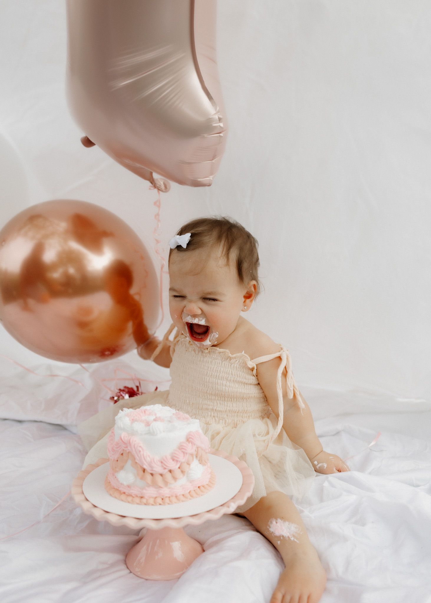A joyful baby laughs during a cake smash photo session with pink and rose gold decorations.