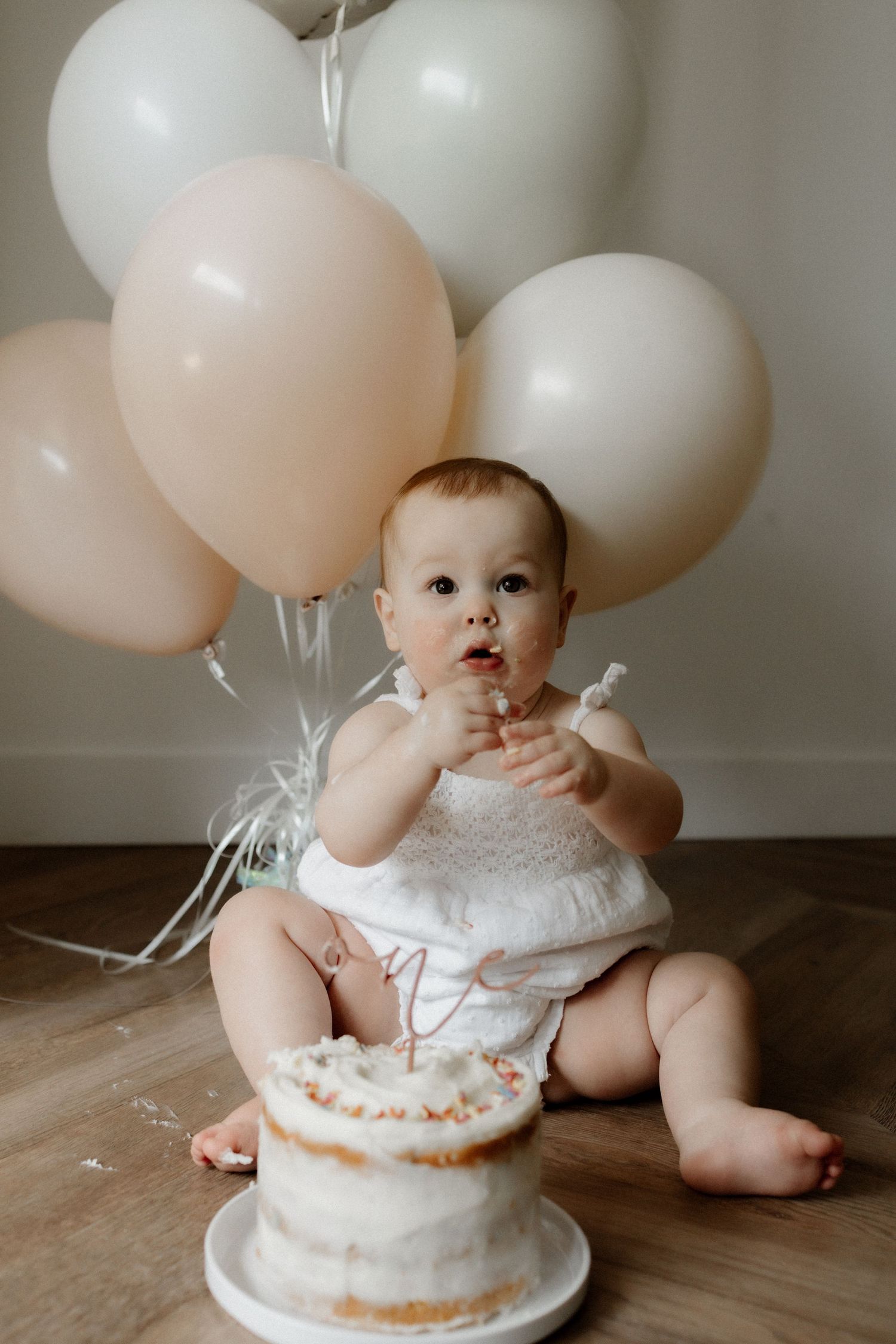 Baby reaching towards white birthday cake with pink and white balloons behind.