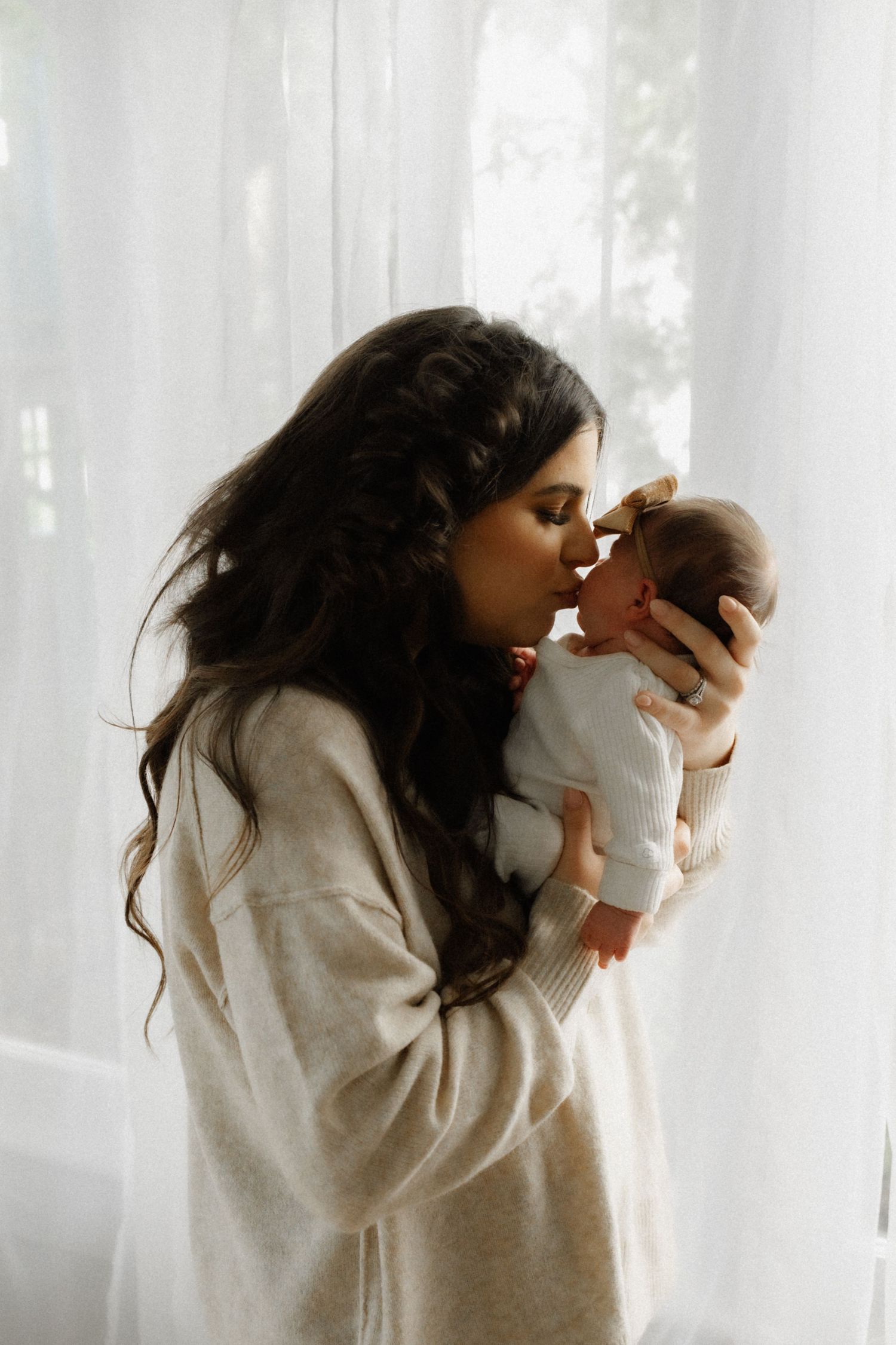 Intimate moment between caregiver and baby against white curtained window backdrop.