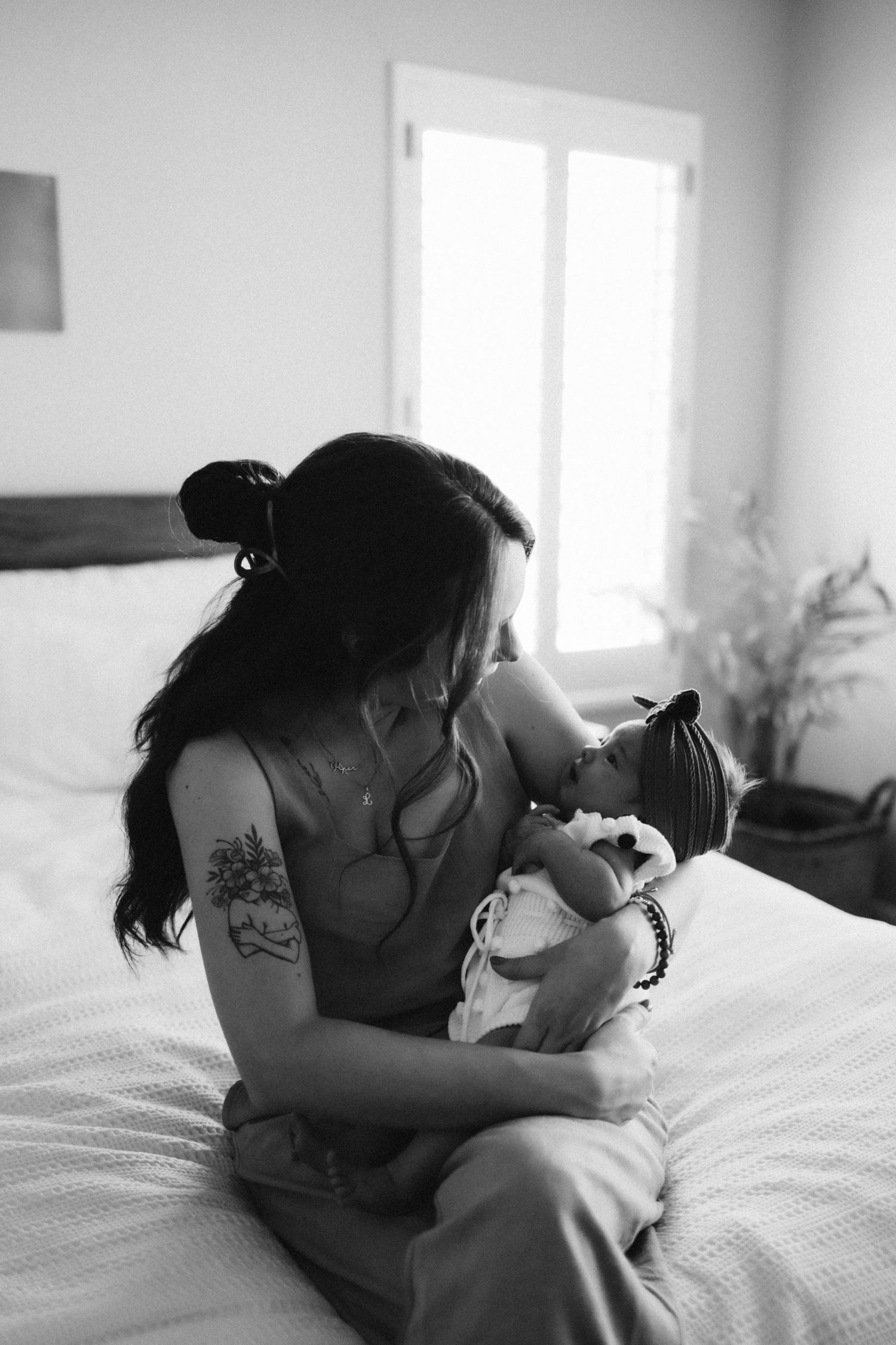 An intimate black and white photo of mother and newborn sharing a tender moment on a bed near a window.