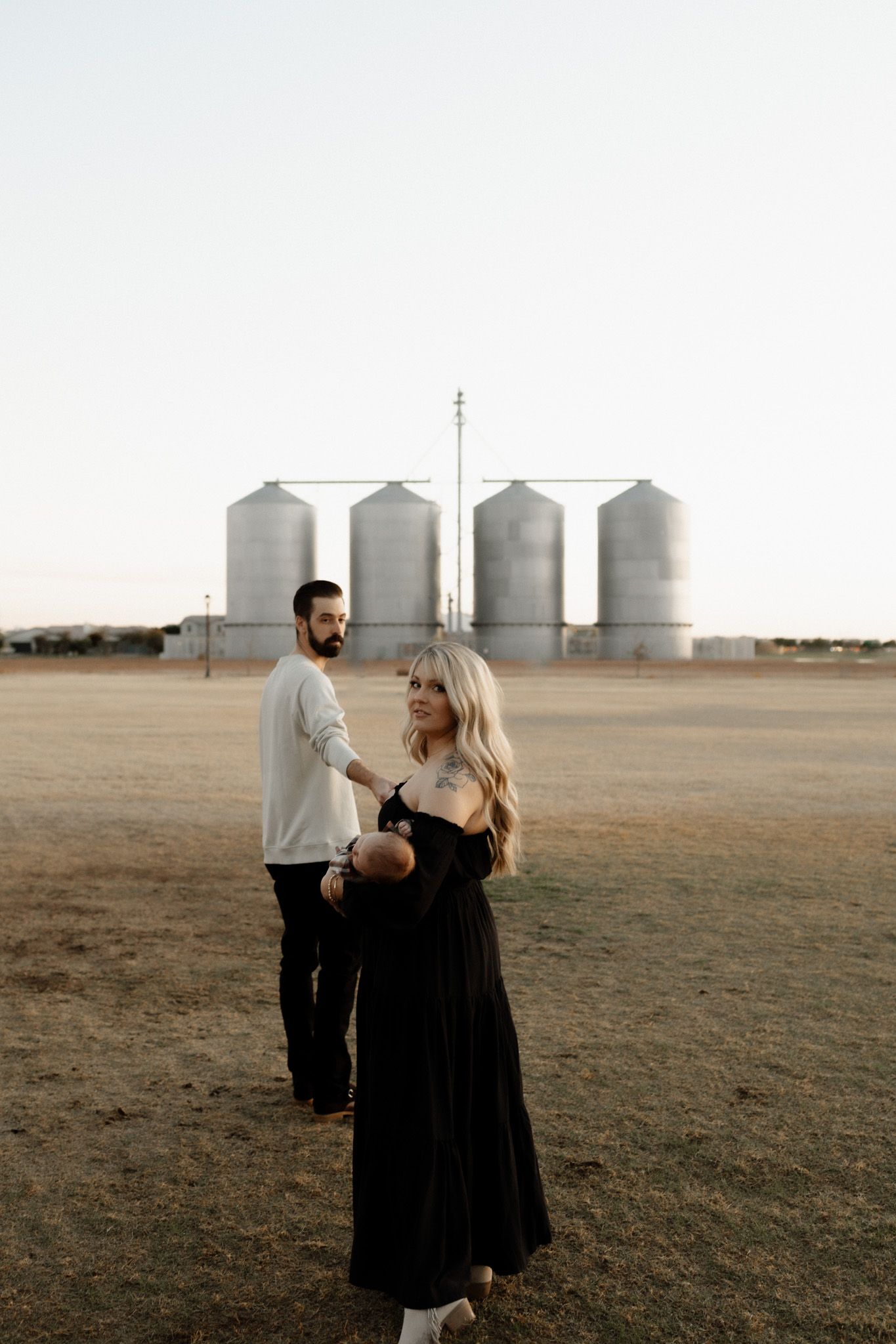 A couple standing in front of large agricultural grain silos at sunset in a rural setting.
