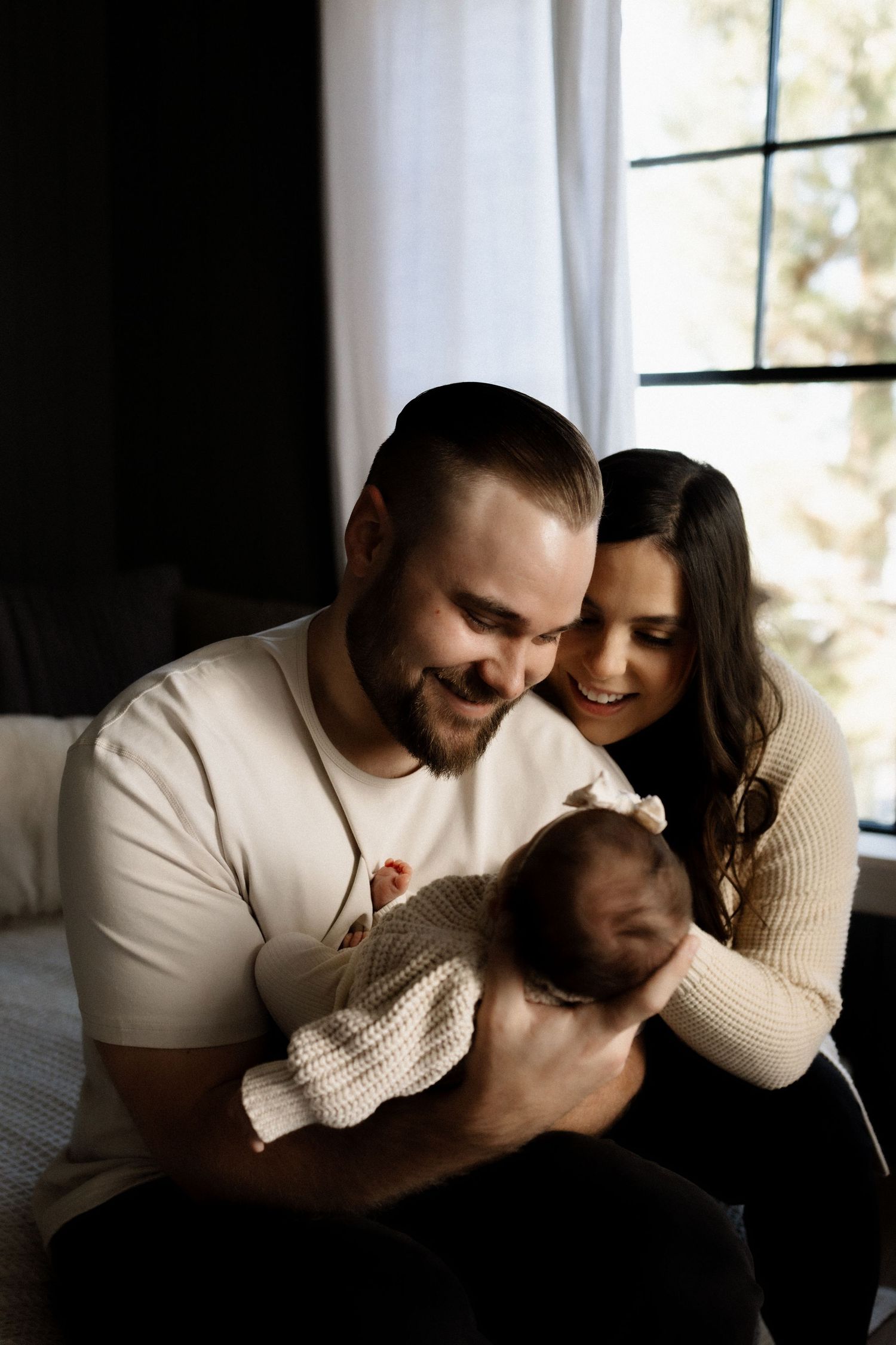 Family shares intimate moment together while holding their newborn in natural window light.