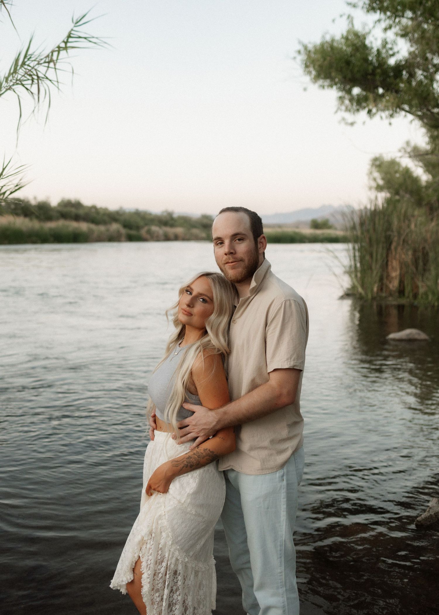 Romantic couple embraces by the waterfront near tall reeds during a serene sunset photoshoot.