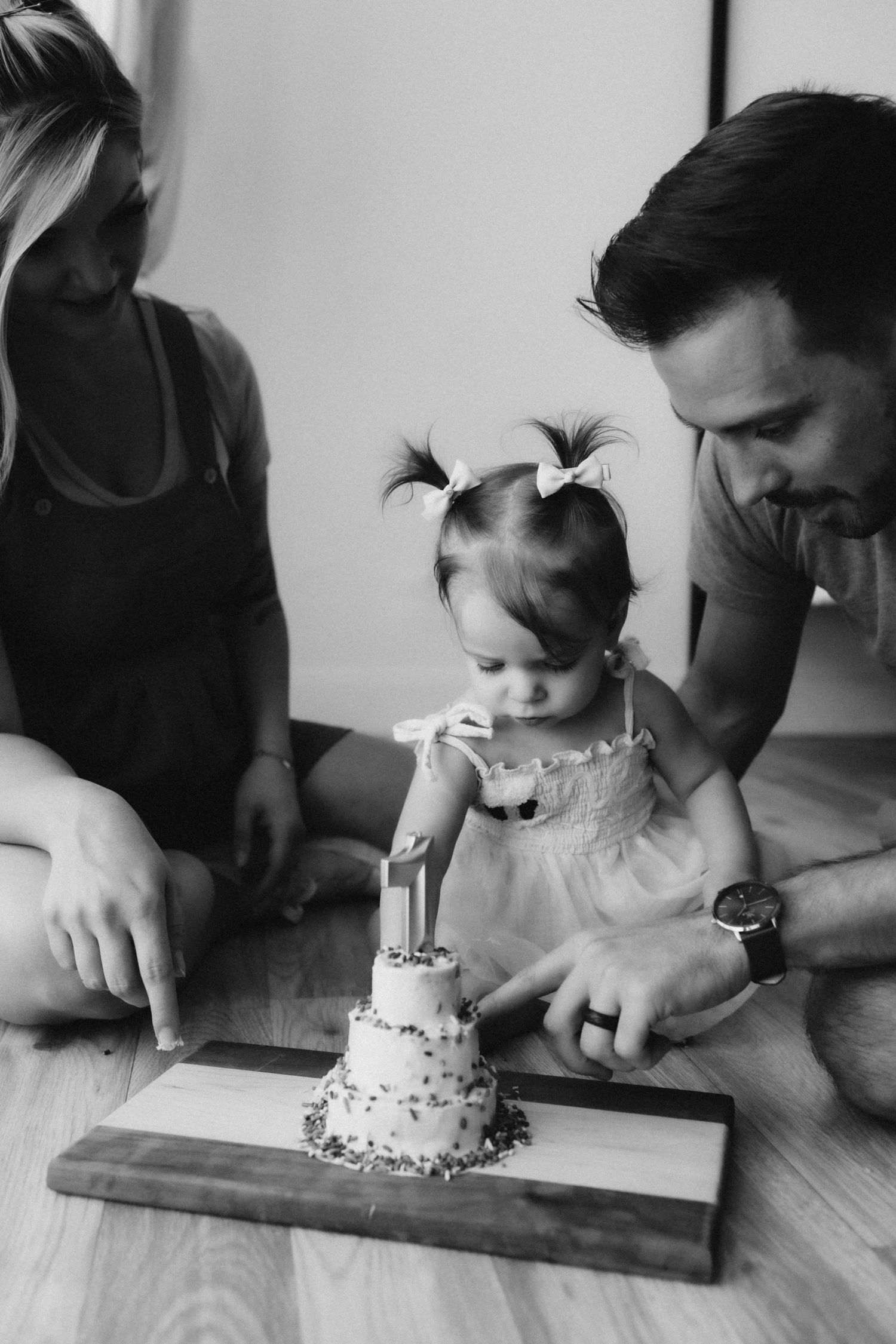Intimate black and white photo of family helping toddler blow out candle on small tiered cake.