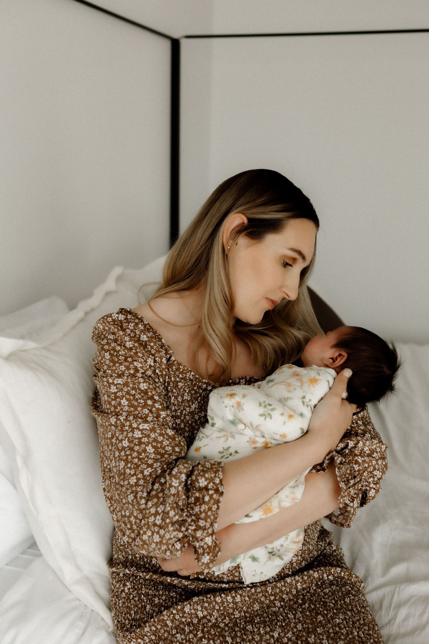 Mother in floral dress cradling newborn baby on white bed.
