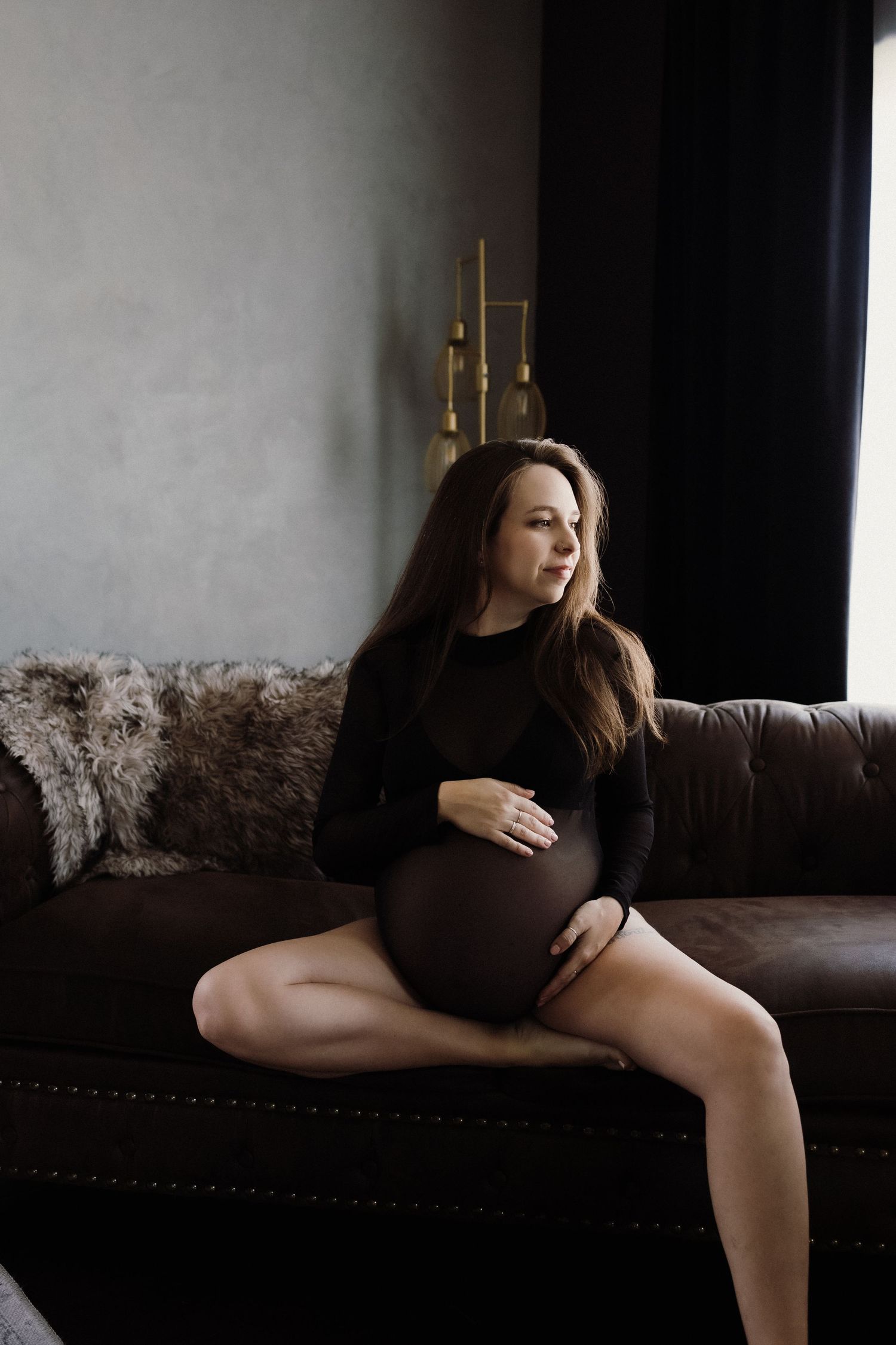 Maternity photo of expectant mother in black dress on leather couch with gold lamp.