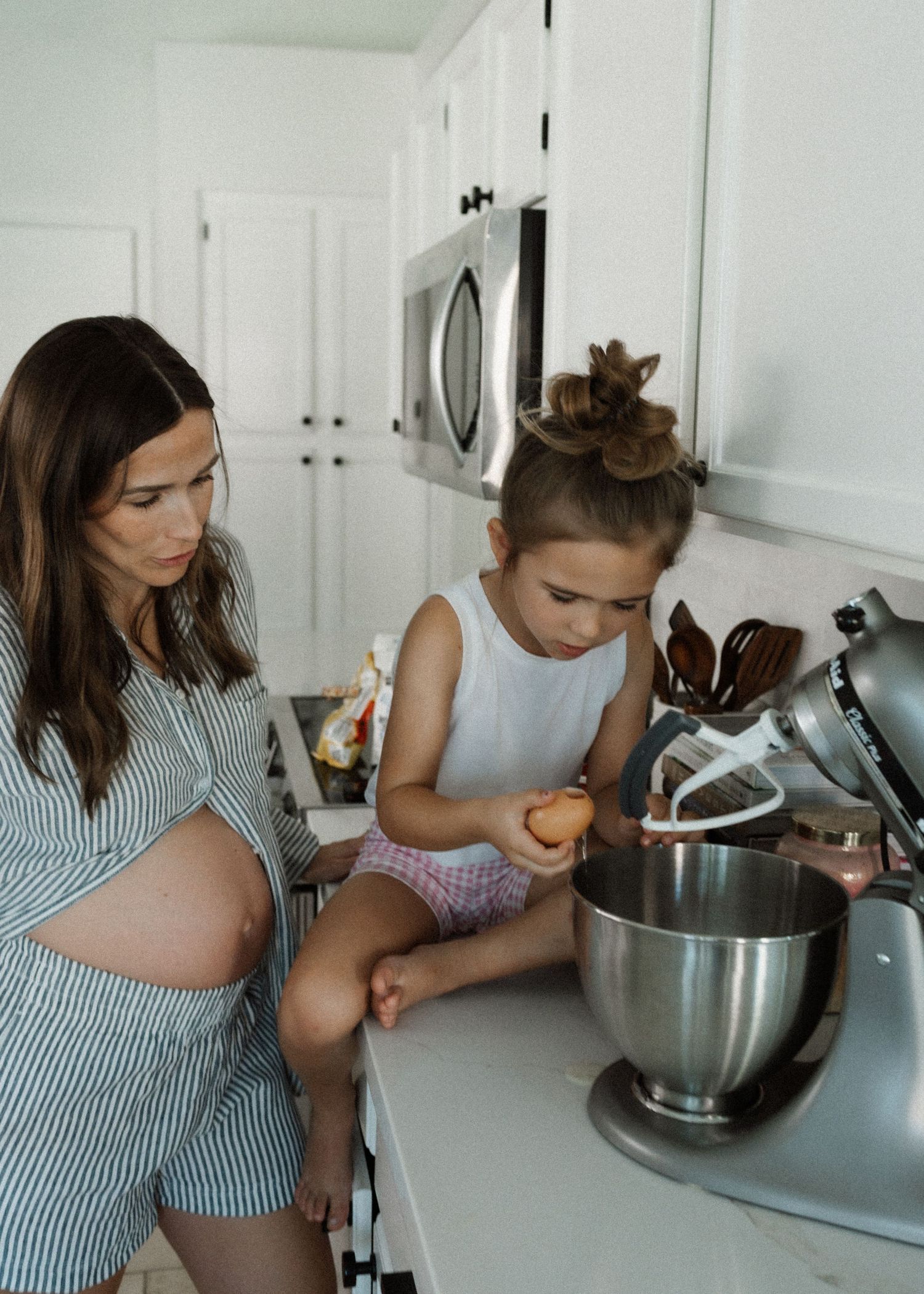A parent and child stand at a kitchen counter working together to mix ingredients in a large silver mixing bowl.
