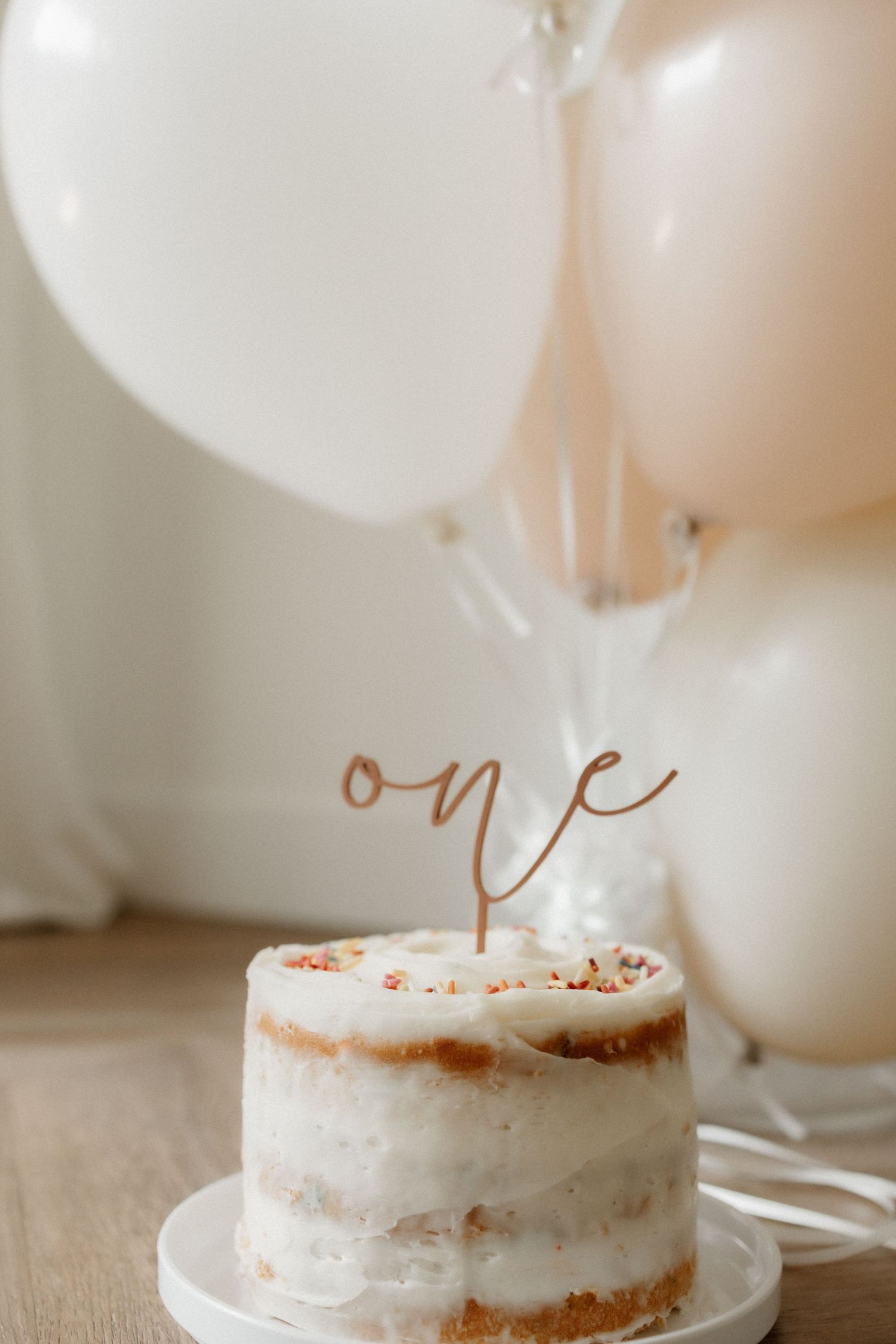 White birthday cake with rose gold 'one' topper against neutral balloons background.