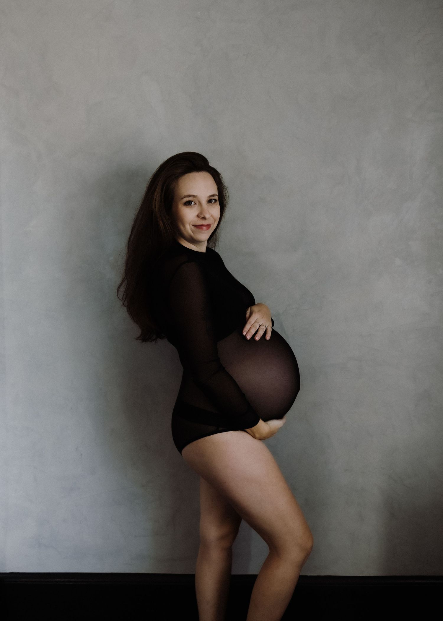 Artistic maternity photo of a person in a black bodysuit against a gray backdrop.