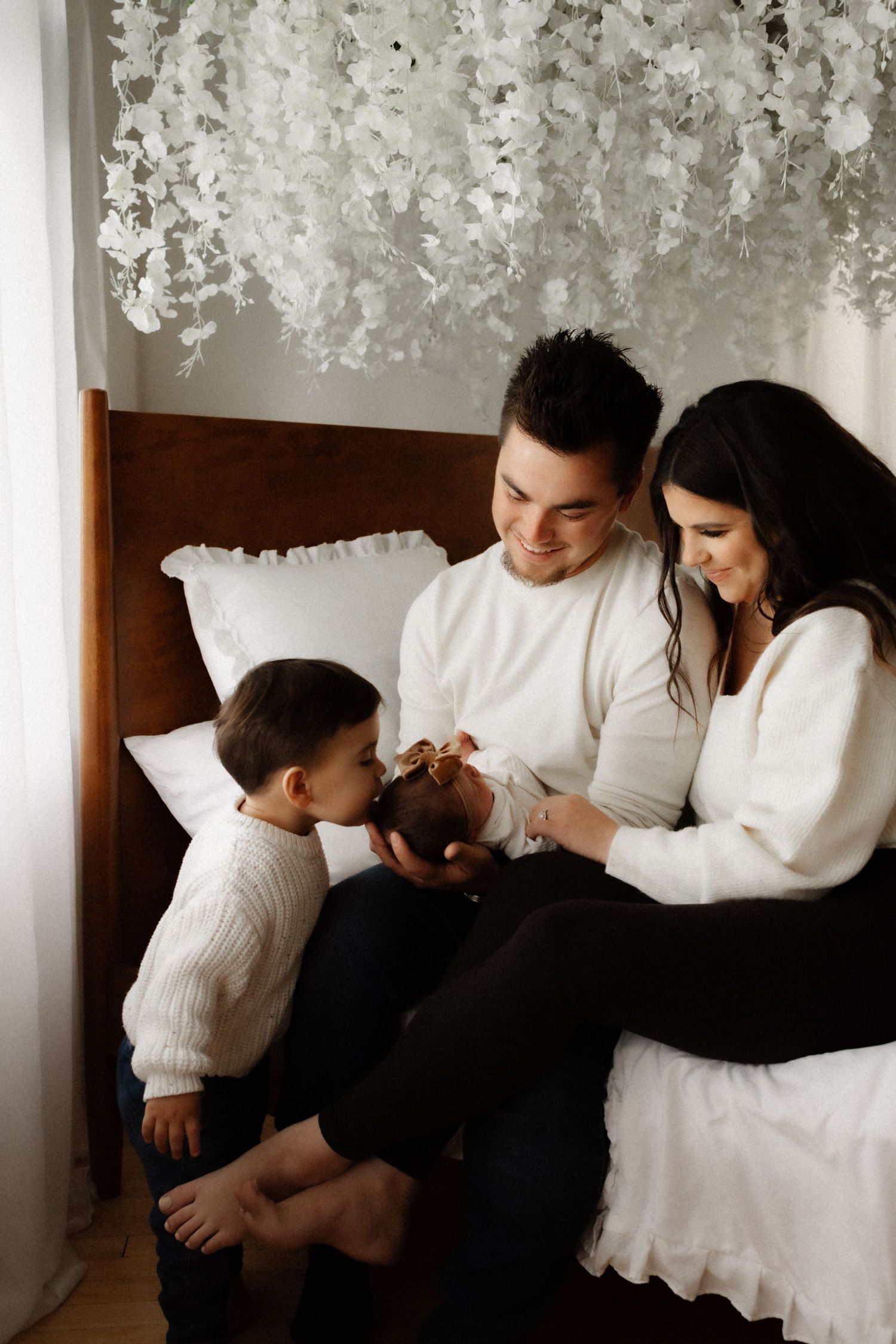 Family in coordinated white outfits gather on bed under hanging floral decorations.