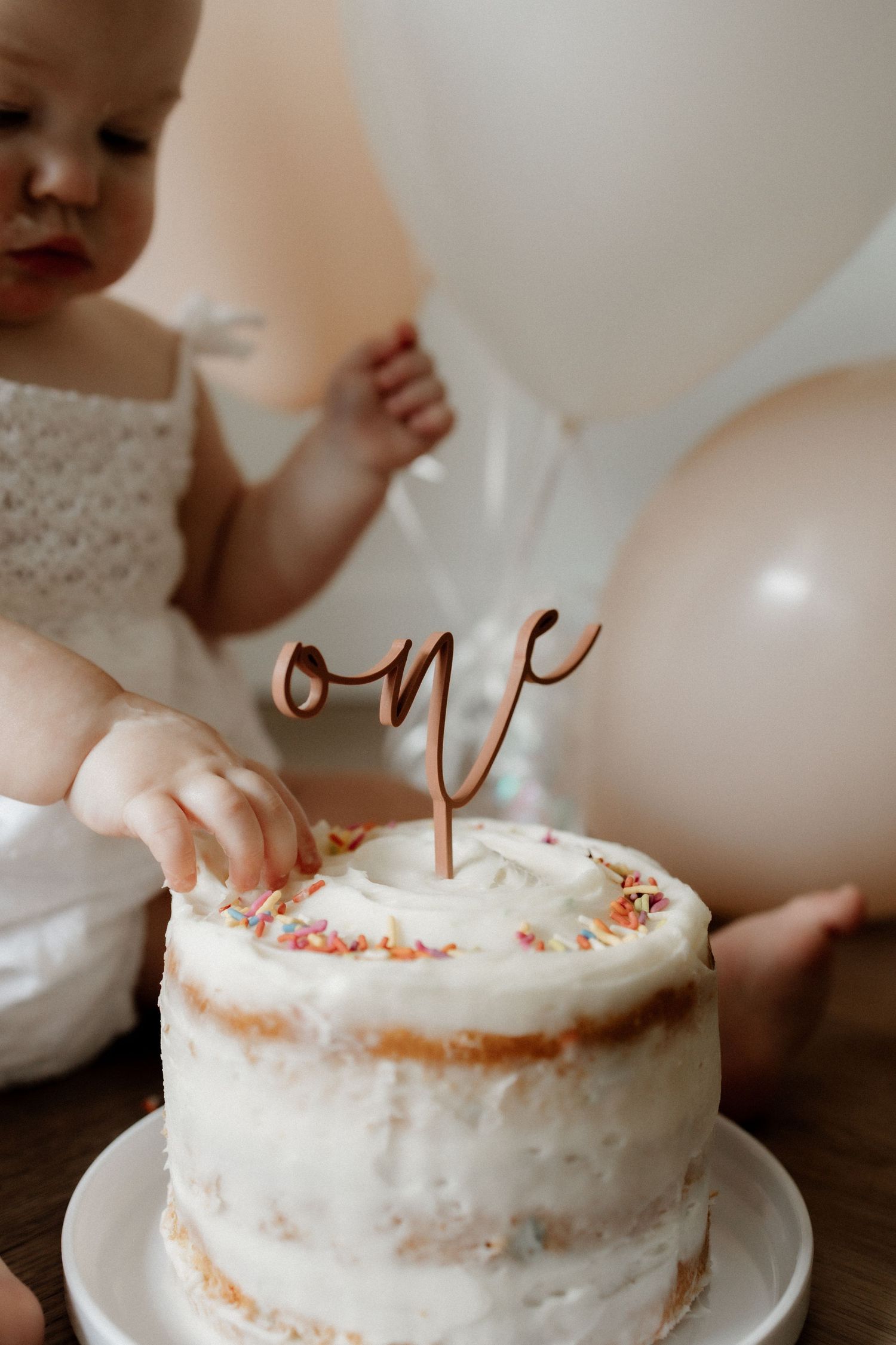 Small white cake with rose gold 'one' topper and sprinkles for first birthday celebration.